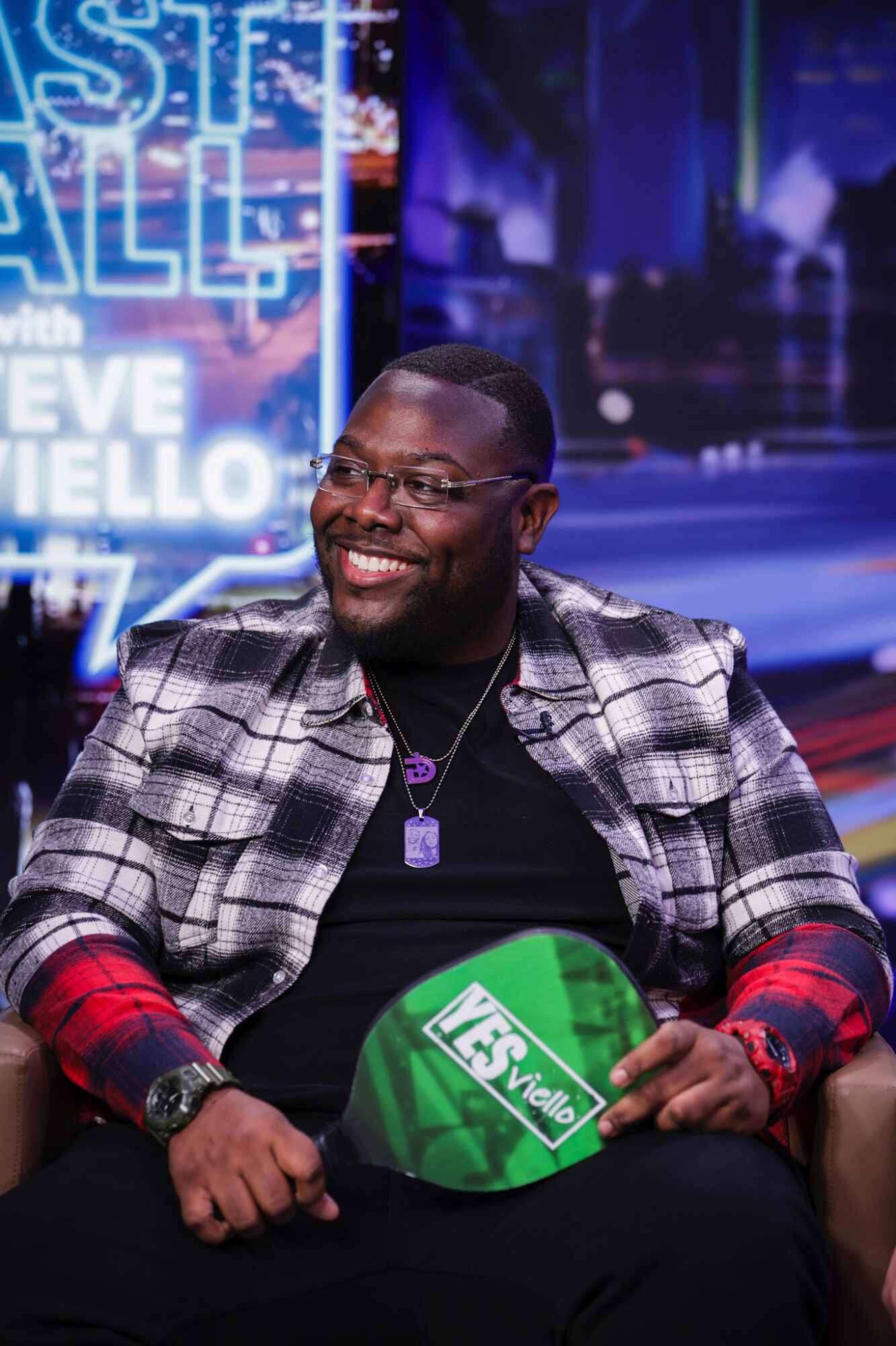 Smiling man with glasses and a checkered jacket, sitting with a green sign, in a TV studio setting.