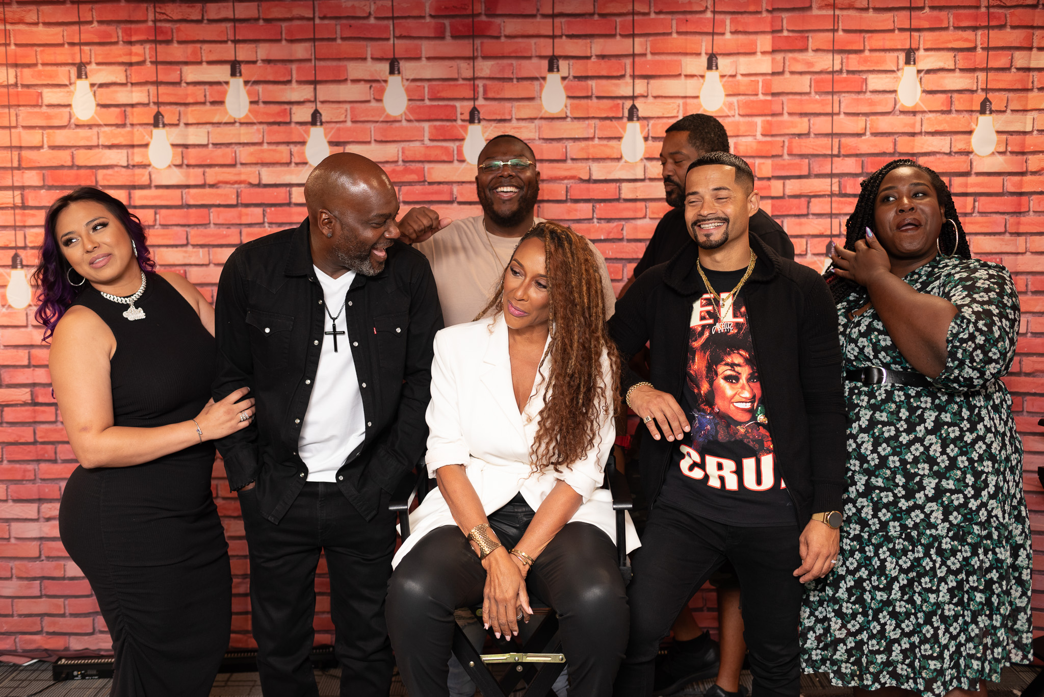 Group of six diverse people smiling and laughing in front of a brick wall with hanging lights.