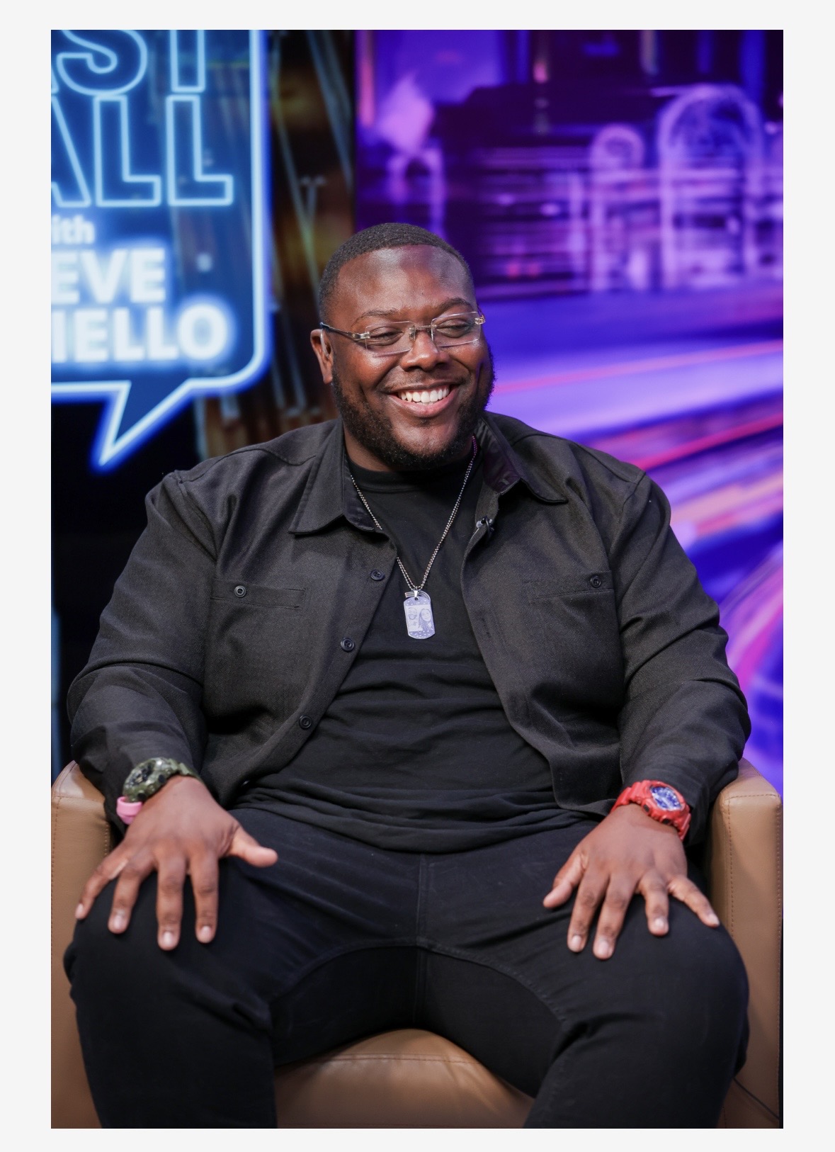Smiling man sitting on a chair, wearing glasses, black shirt, and a necklace, in a studio setting with neon signs.