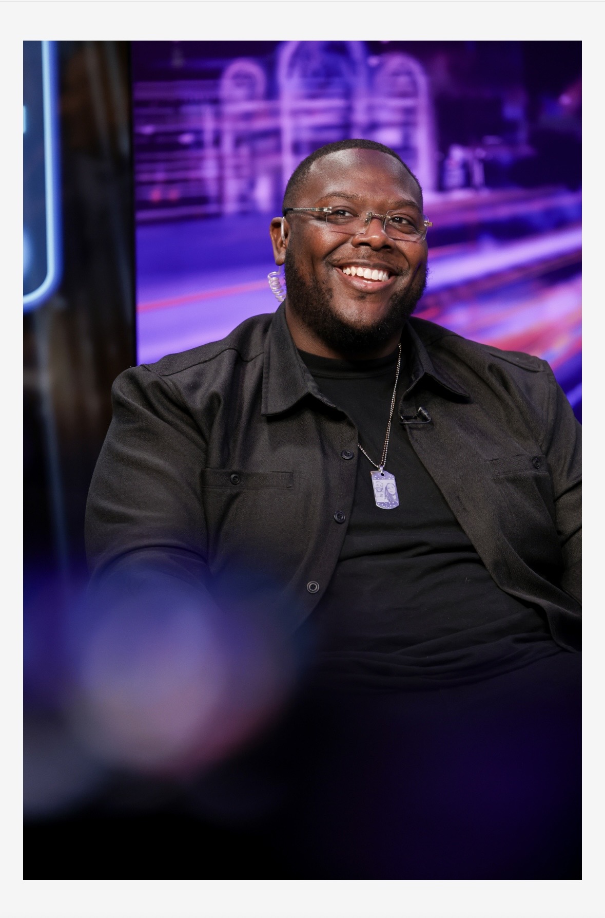 Smiling man with glasses and a necklace, sitting indoors with purple and blue neon lights in background.