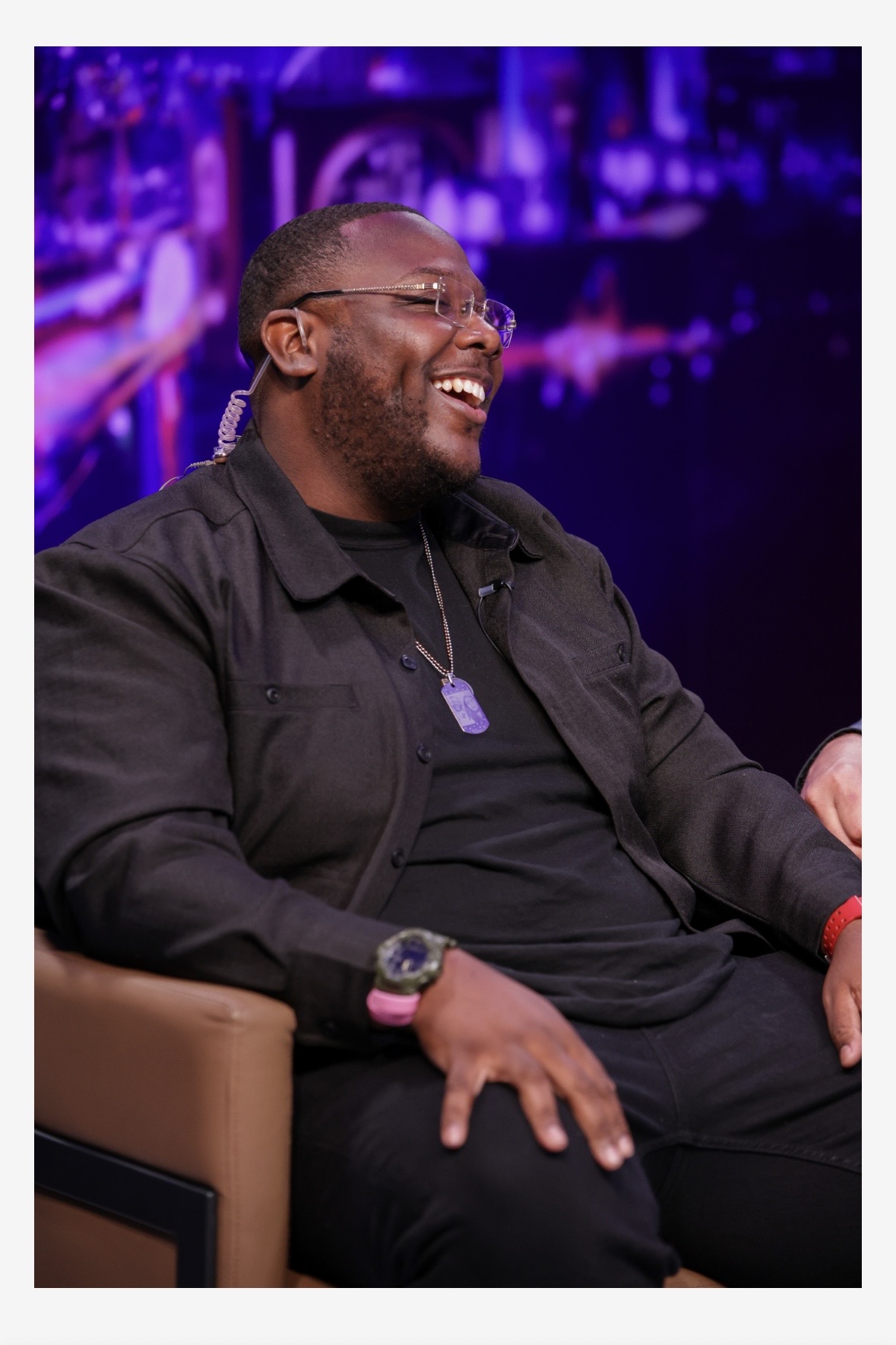 Man with glasses and beard smiling, sitting in a chair with purple lighting background.