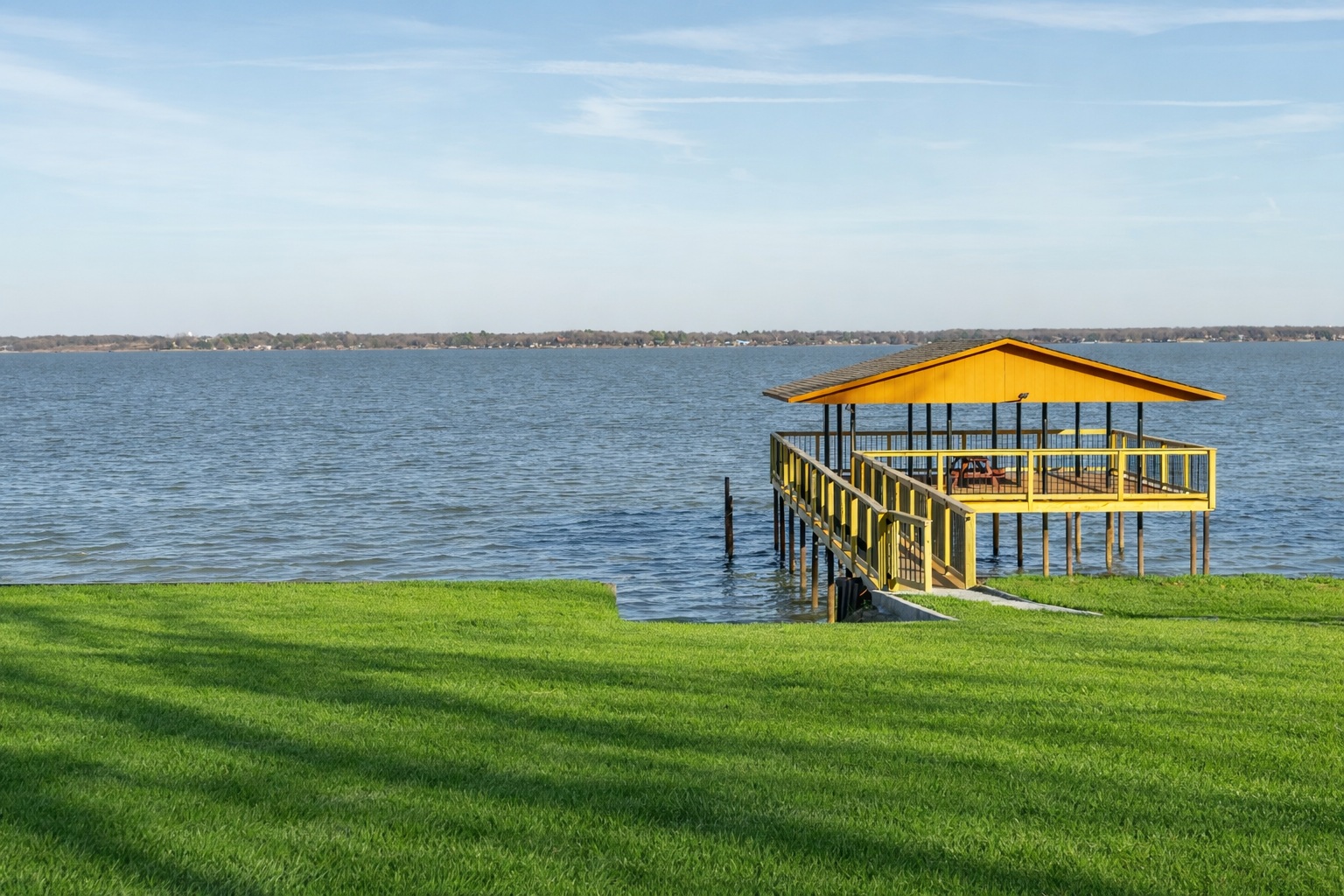Yellow pavilion on stilts over water with green grass in foreground and blue sky.