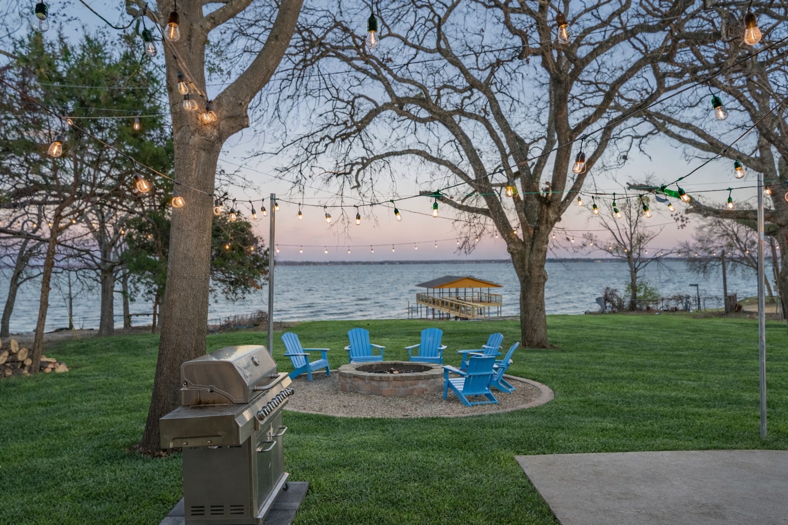 Outdoor scene with trees, string lights, a picnic table, and chairs near water, with a grill in the foreground.