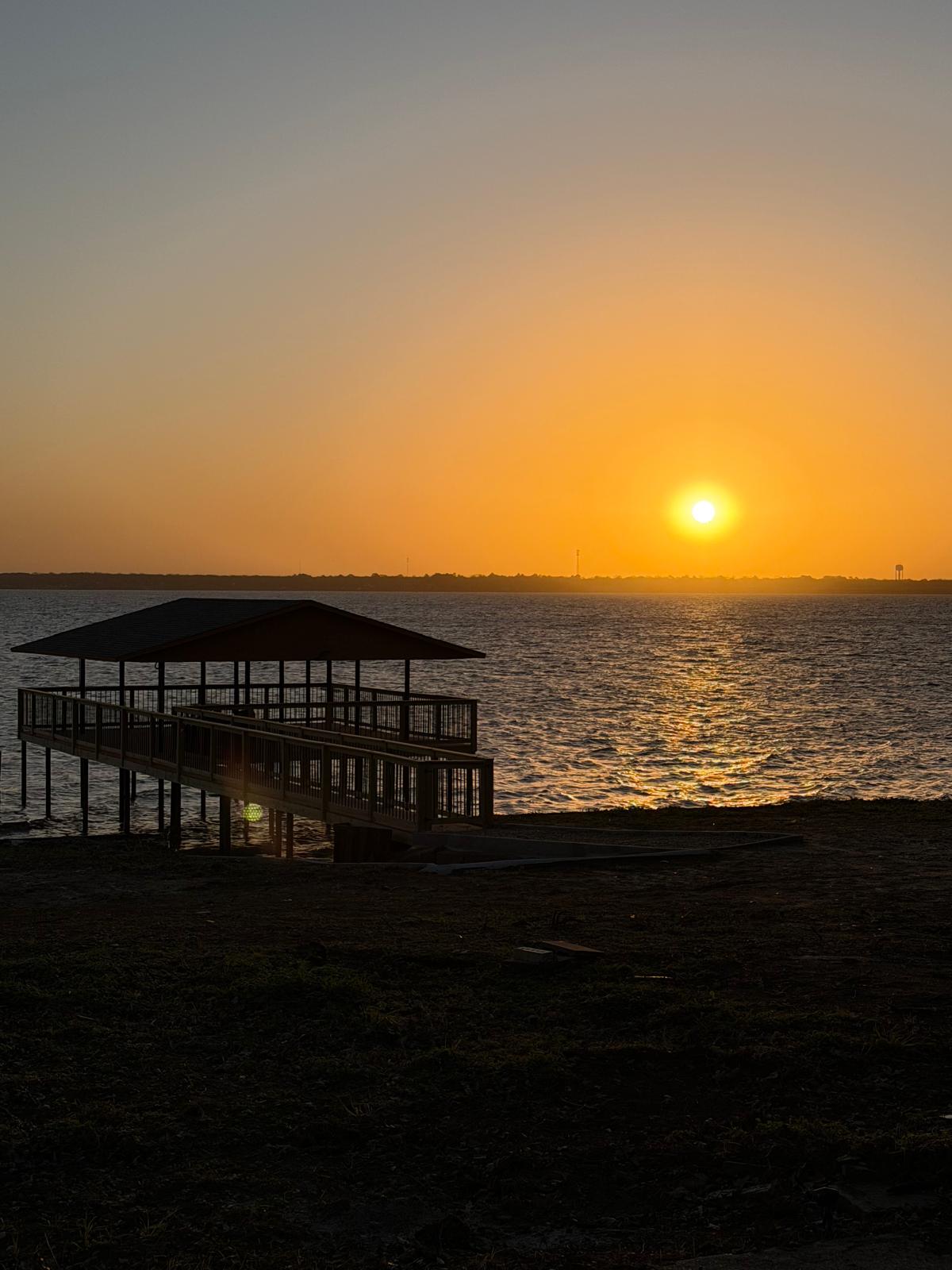 Sunset over water with a pier and shelter in silhouette, sky transitioning from orange to blue.