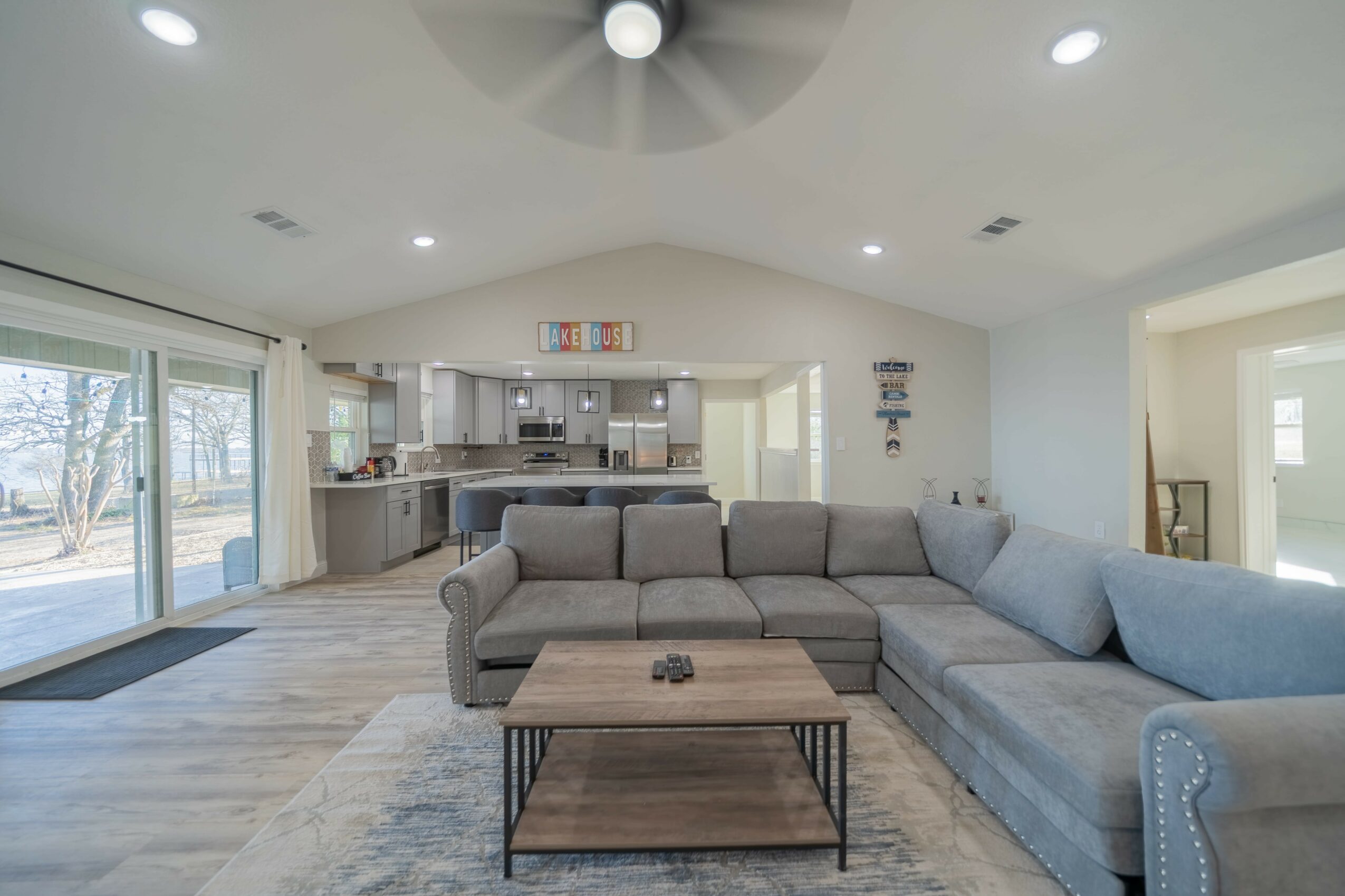 Living room with gray sectional sofa, wooden coffee table, open kitchen, and sliding glass door to outside.