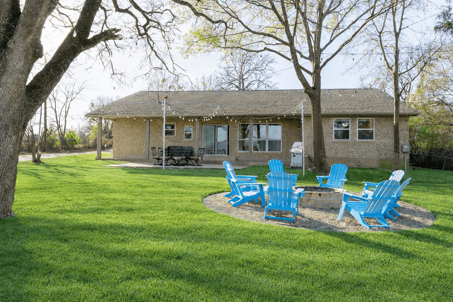 Backyard with a house, trees, and a circle of blue chairs around a fire pit on grass.