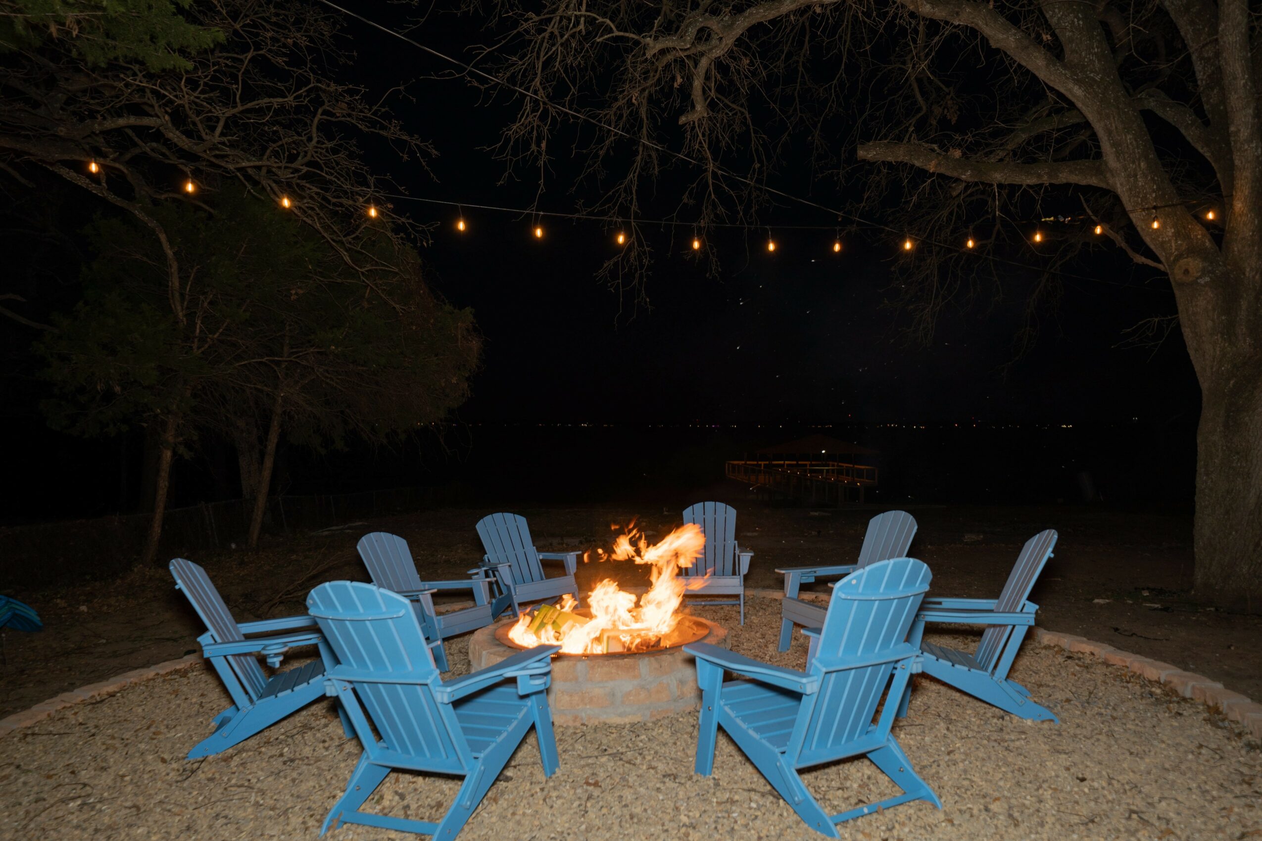 Eight blue Adirondack chairs arranged in a circle around a campfire at night, with string lights overhead and trees in background.