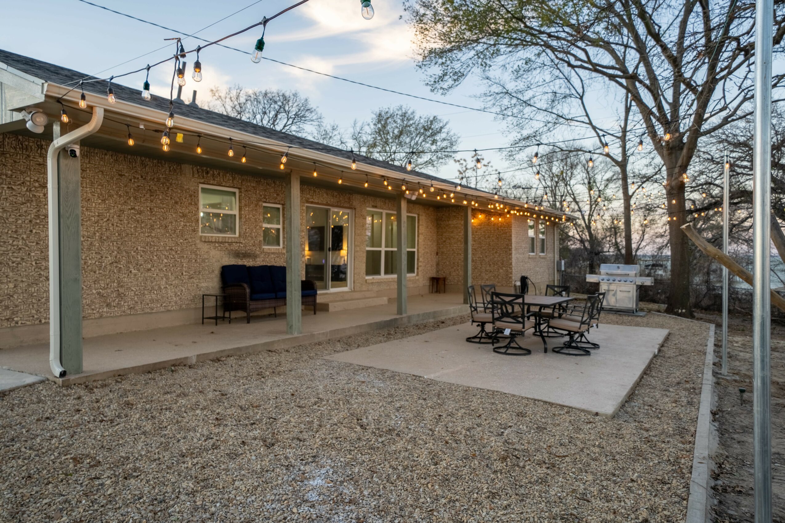 Backyard patio with string lights, outdoor furniture, and a concrete area, surrounded by trees and a house wall.