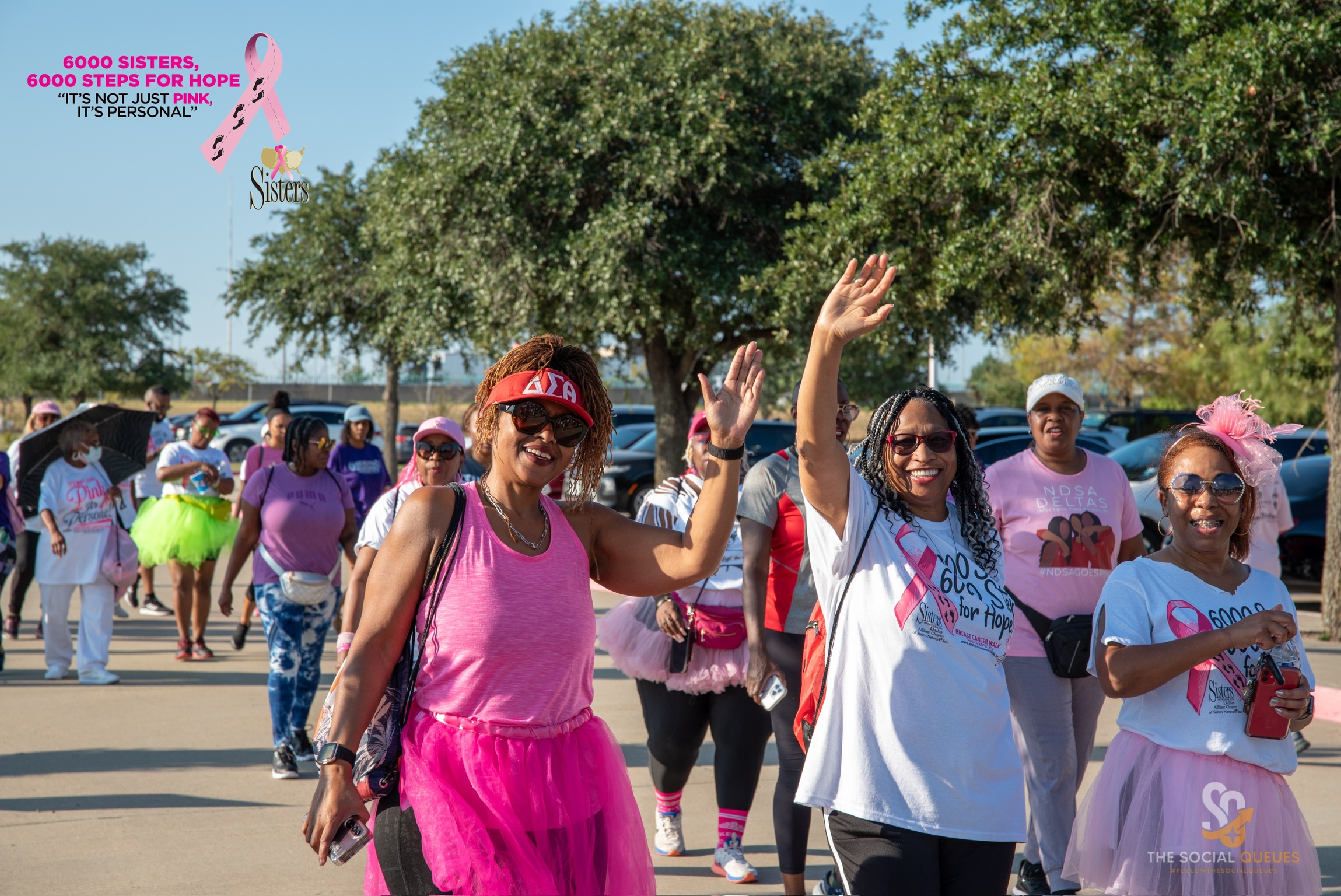 Group of women participating in a walk event outdoors, some waving and smiling, trees and parked cars in background.