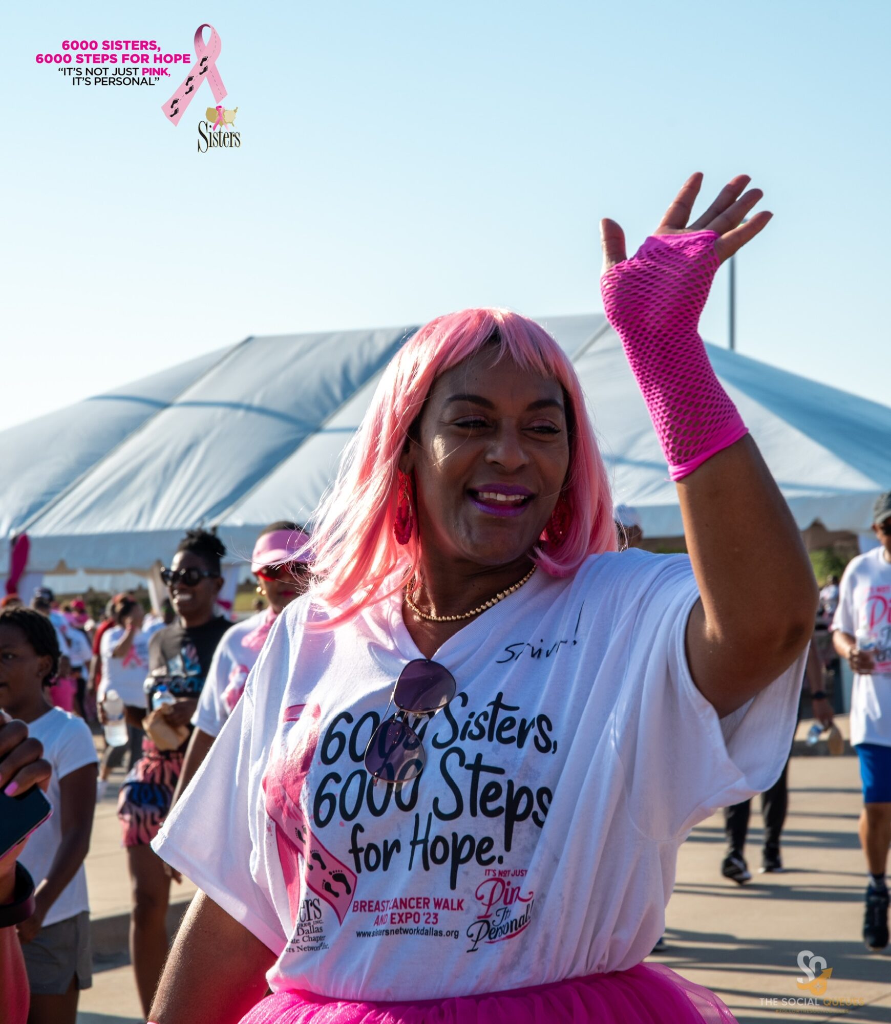 Smiling woman with pink hair waving at outdoor event, wearing a white T-shirt with blue and pink text, surrounded by people.