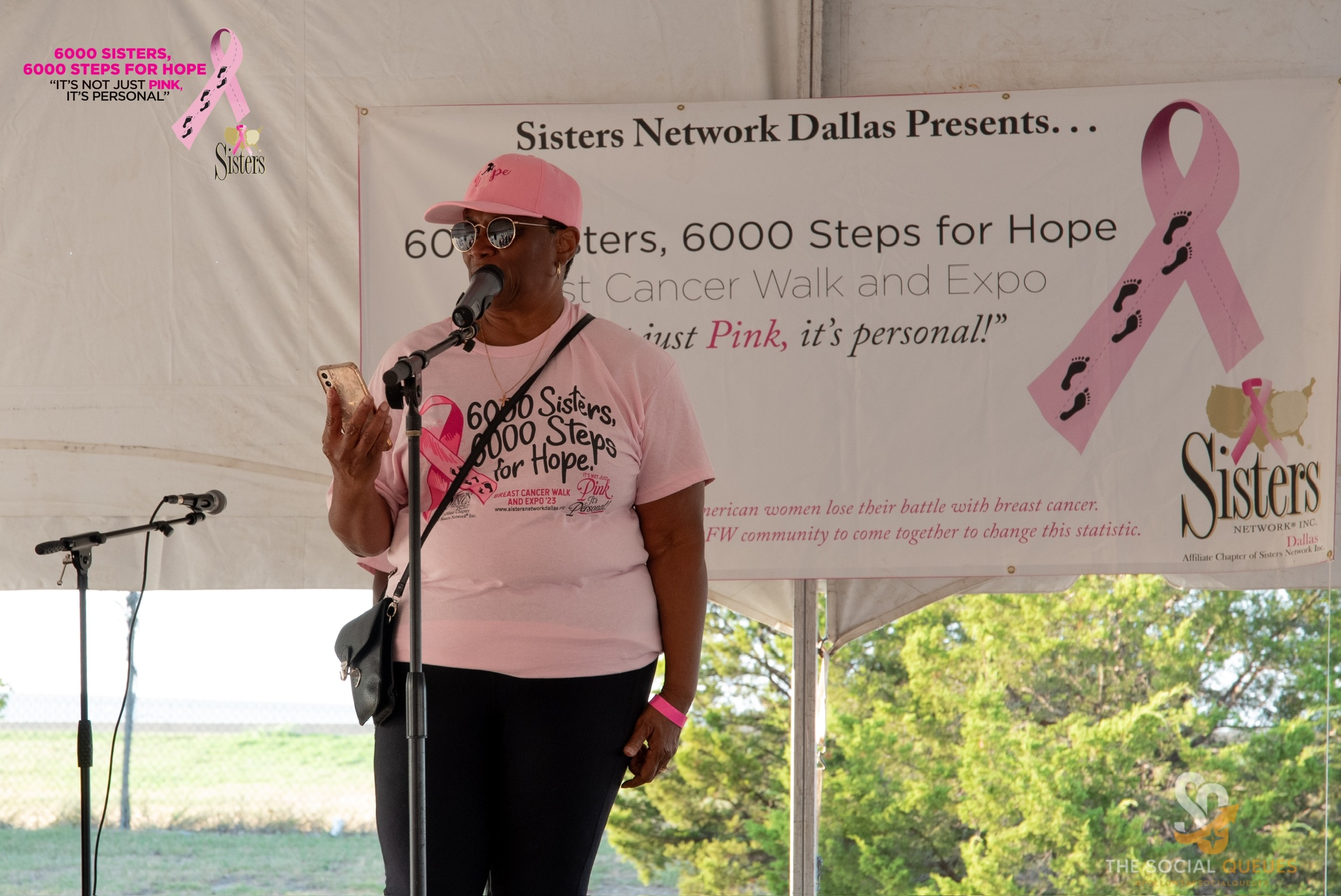 Woman speaking into microphone at breast cancer awareness event, pink ribbon banner behind her, outdoors setting.