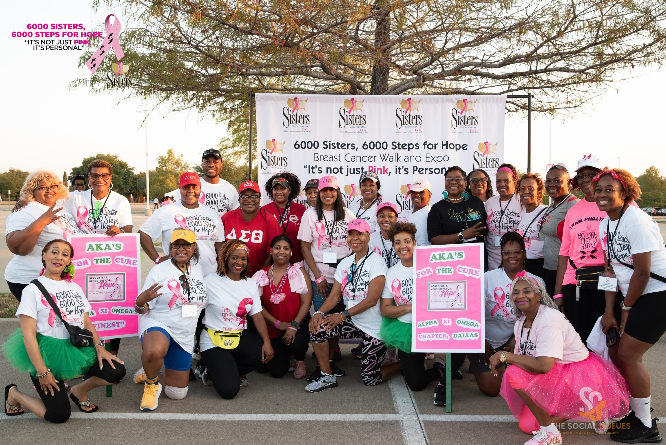 Group of people participating in breast cancer awareness walk, holding pink signs and banners, outdoors with trees in background.