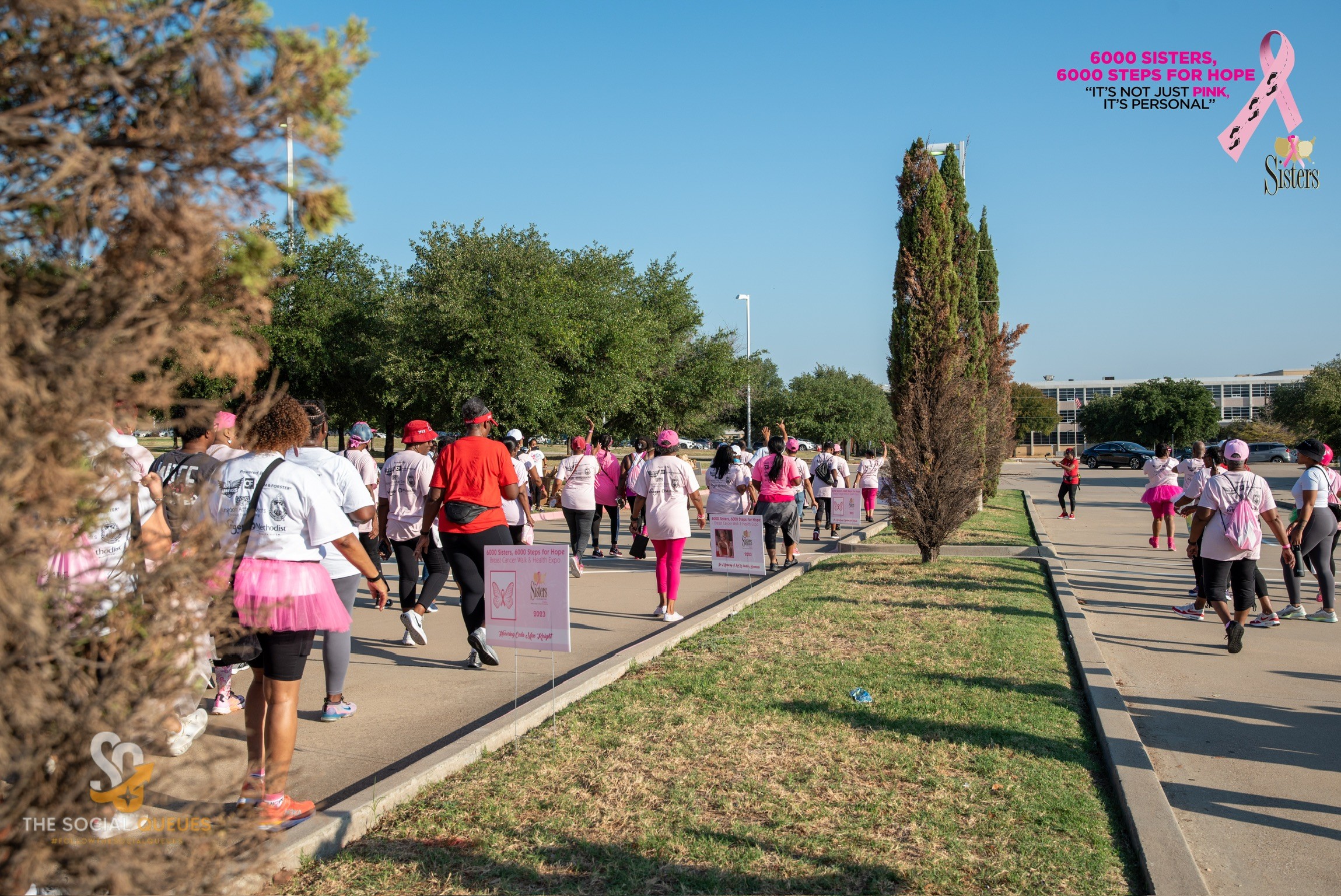 Group of people walking outdoors on a sunny day, some wearing pink skirts and white shirts, with trees and a grassy area.