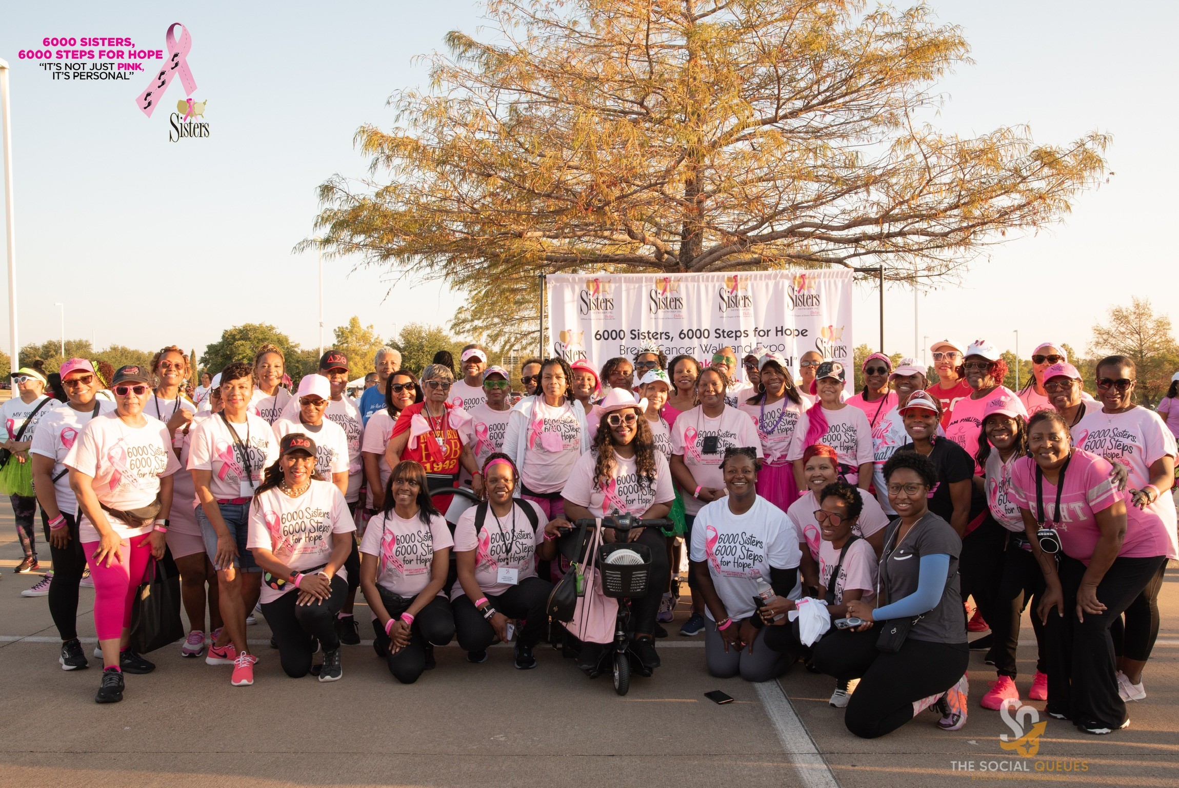 Group of people wearing pink and white shirts gathered outdoors for a walk or event, with a tree and banner in background.
