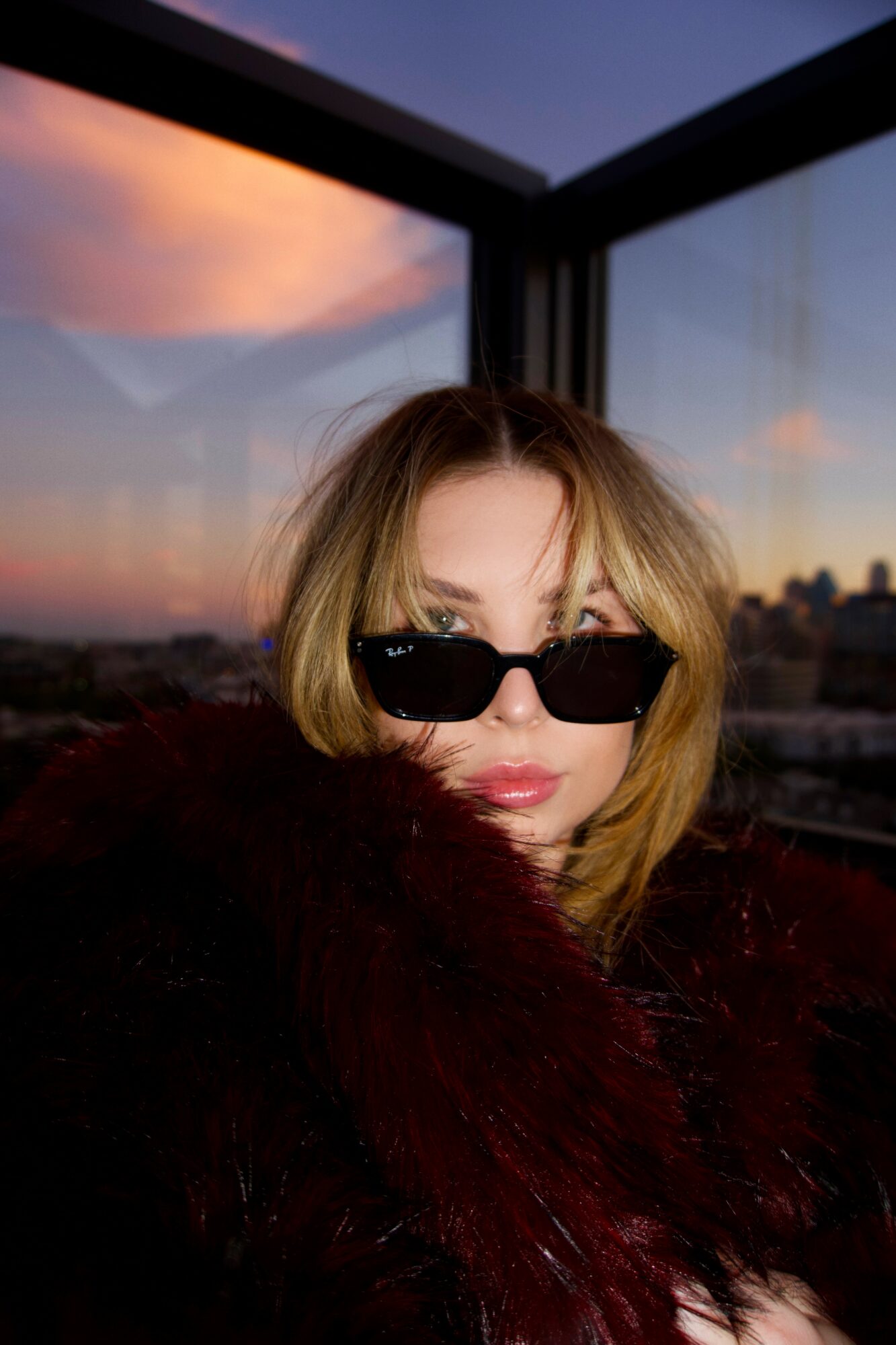 Woman with blonde hair wearing sunglasses, looking at camera, indoors with sunset view outside window.