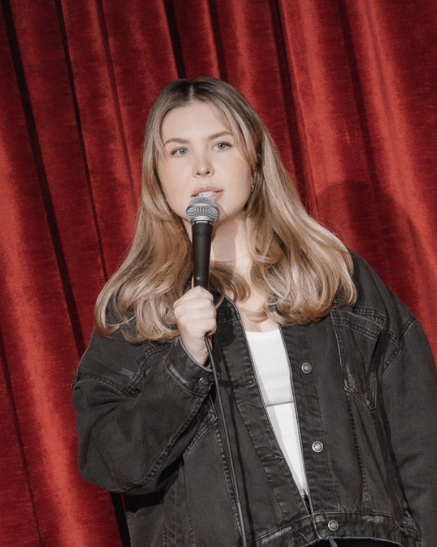 Young woman with long hair holding a microphone, standing in front of red curtains, wearing a black jacket and white shirt.