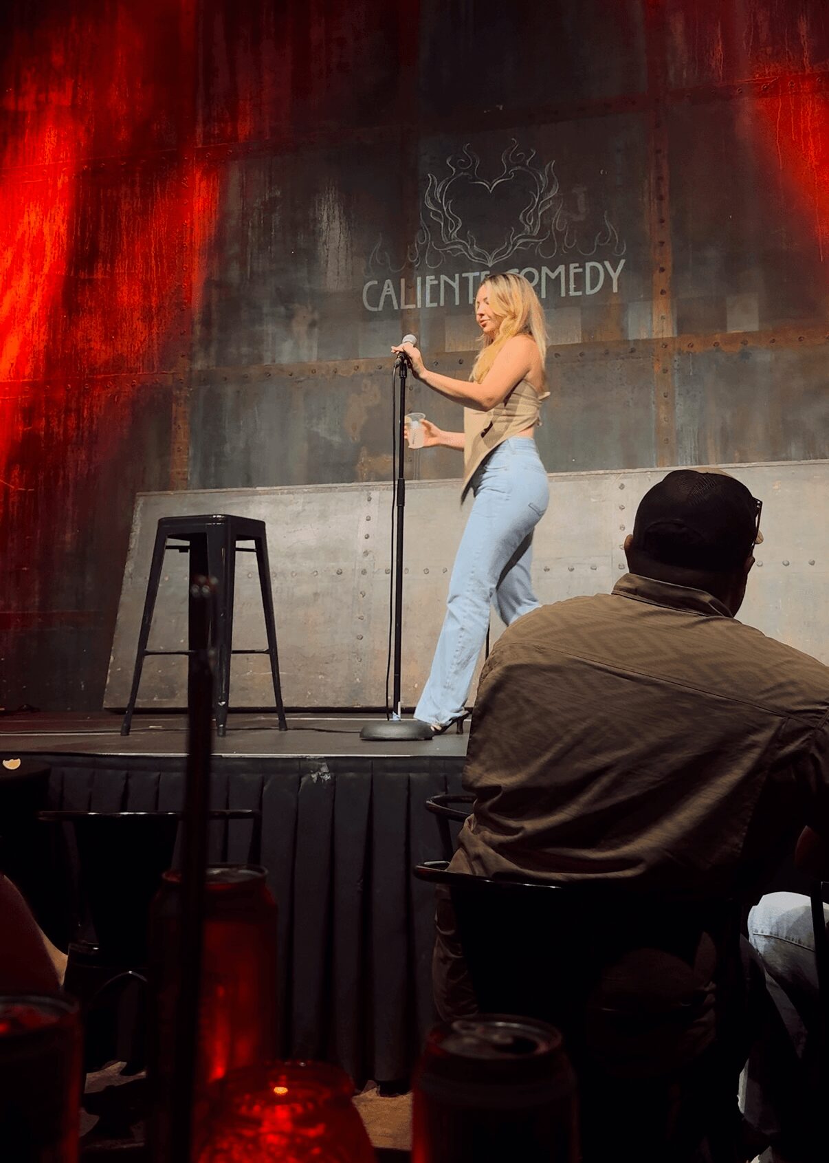 Performer on stage with microphone, woman in jeans, audience member in foreground, stage backdrop with text, stool nearby.