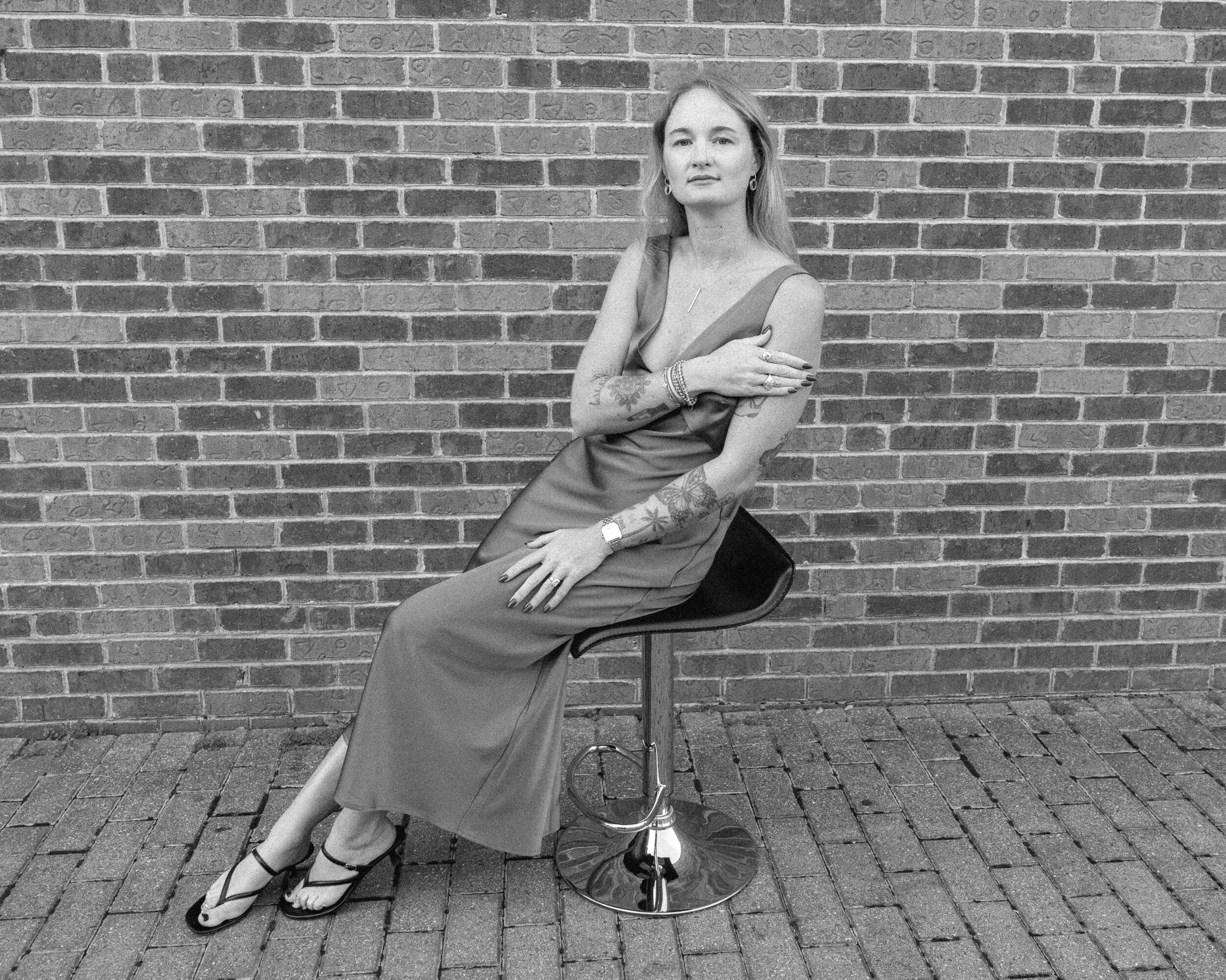 Woman sitting on a stool in front of a brick wall, wearing a long dress and sandals, with arms crossed.