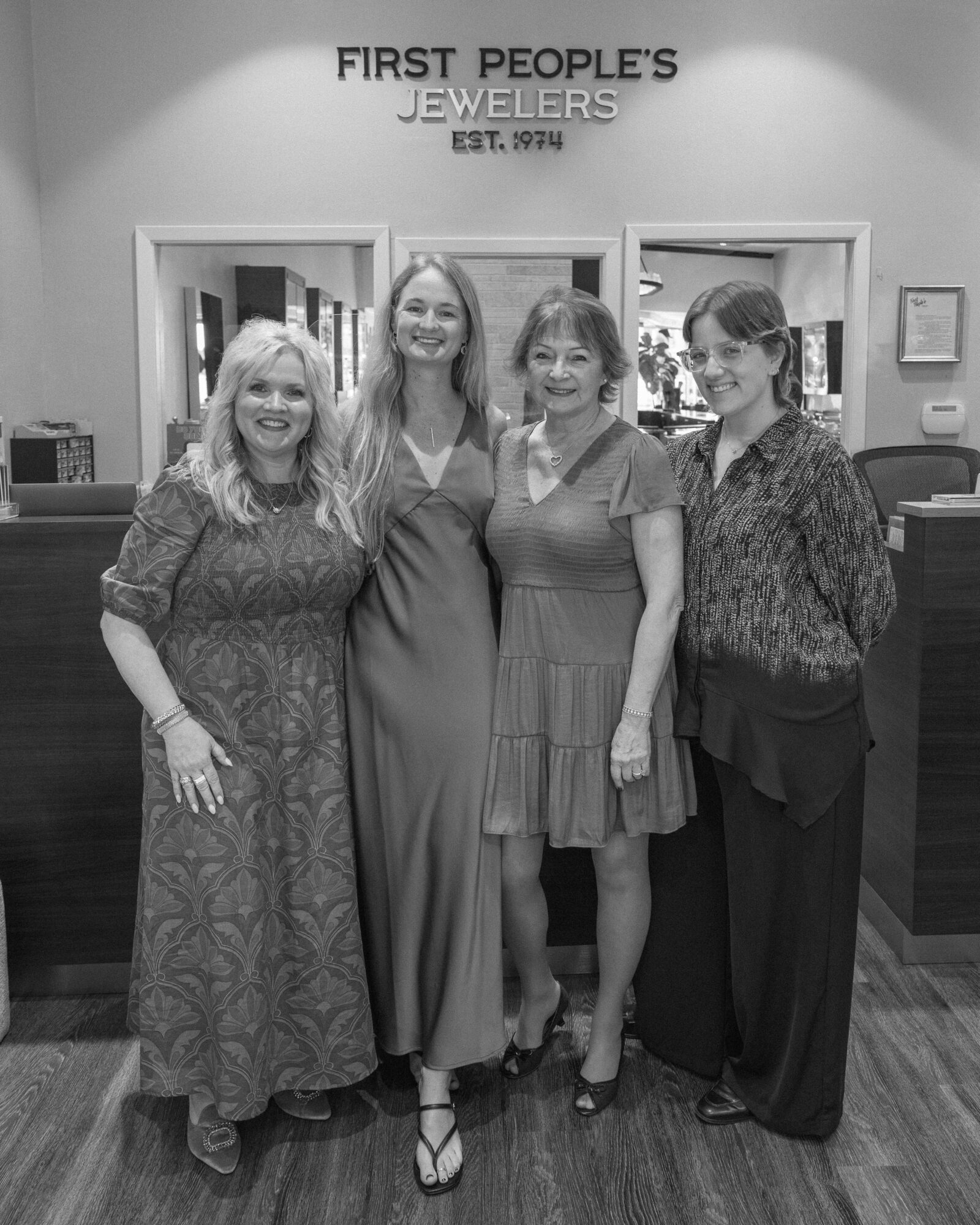 Four women standing in front of a jewelry store, smiling, with a sign above reading 'First People's Jewelers'.