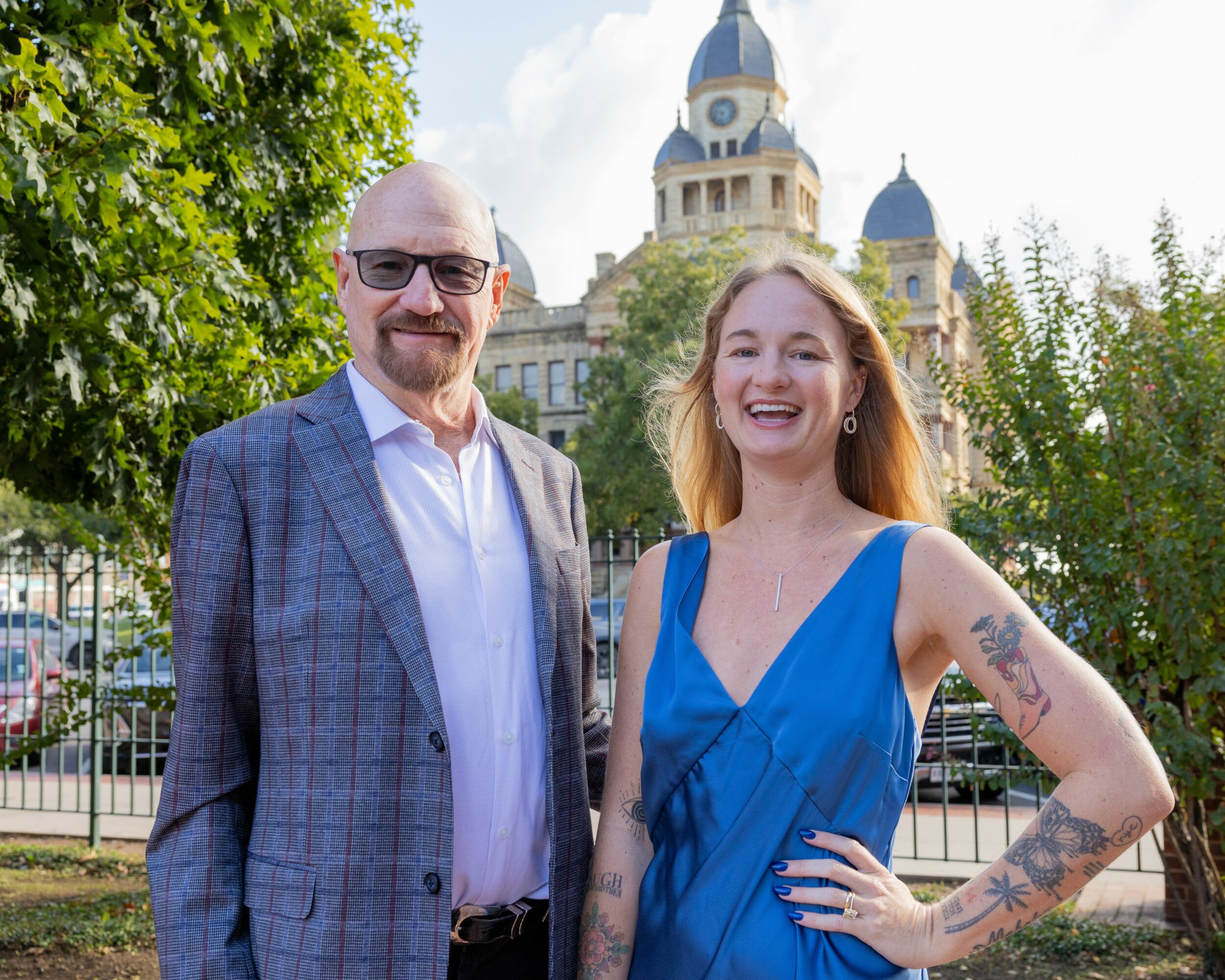 Man in glasses and a woman in a blue dress smiling outdoors with a historic building in background.