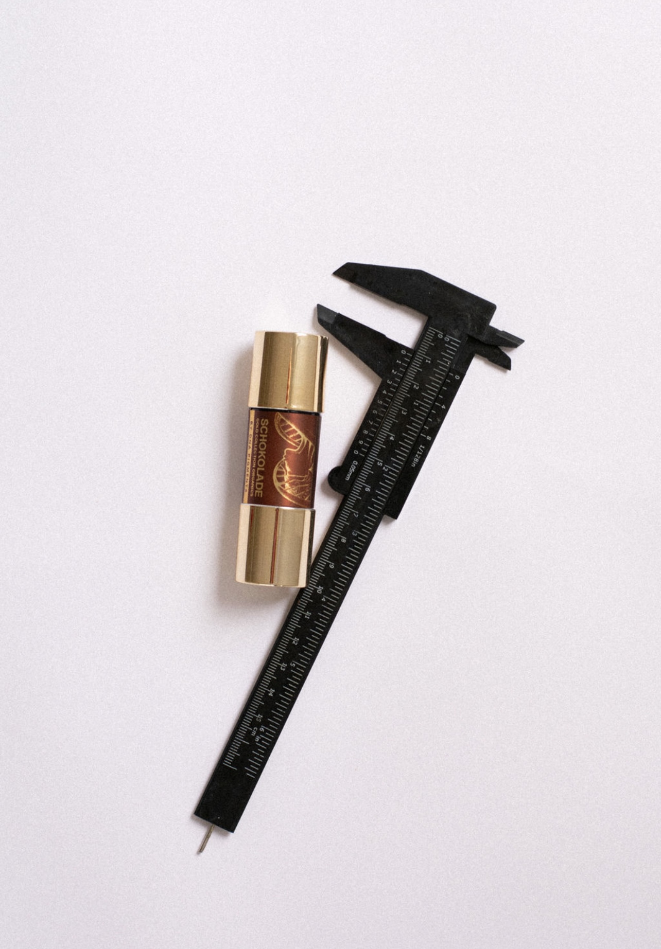 Black caliper measuring a lipstick with gold and brown packaging on a white background.