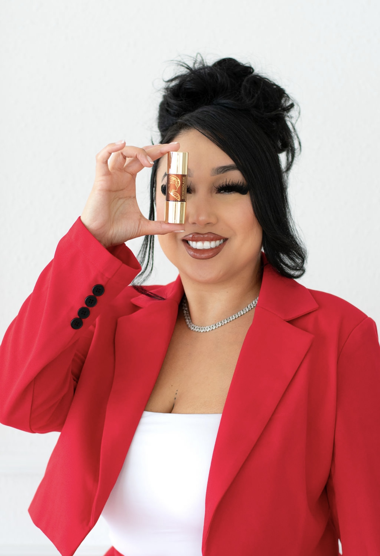 Woman with dark hair in a bun, smiling, holding a small bottle near her eye, wearing a red blazer and white top.