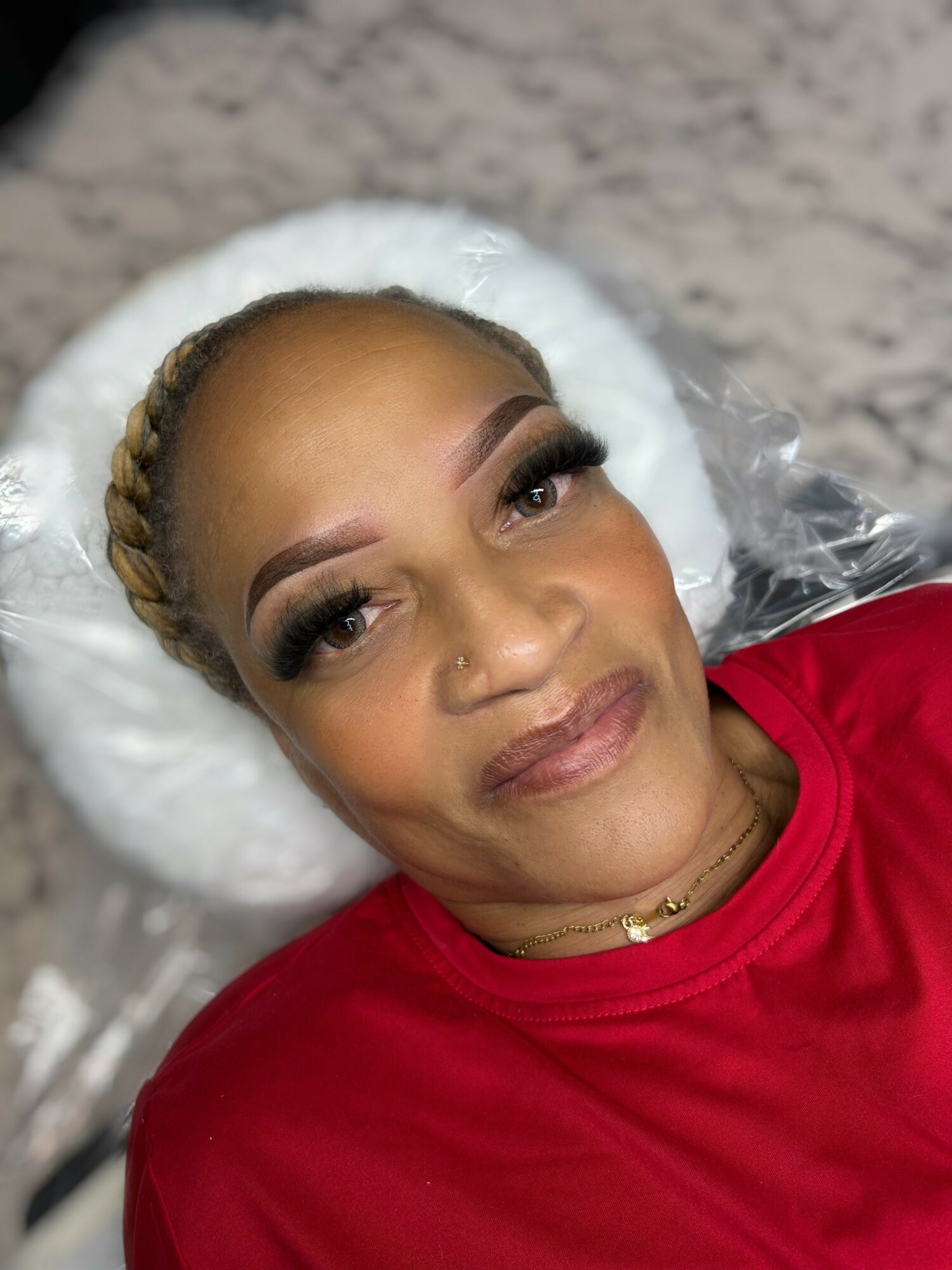 Woman with braided hair and makeup lying on a pillow, wearing a red top, smiling gently.