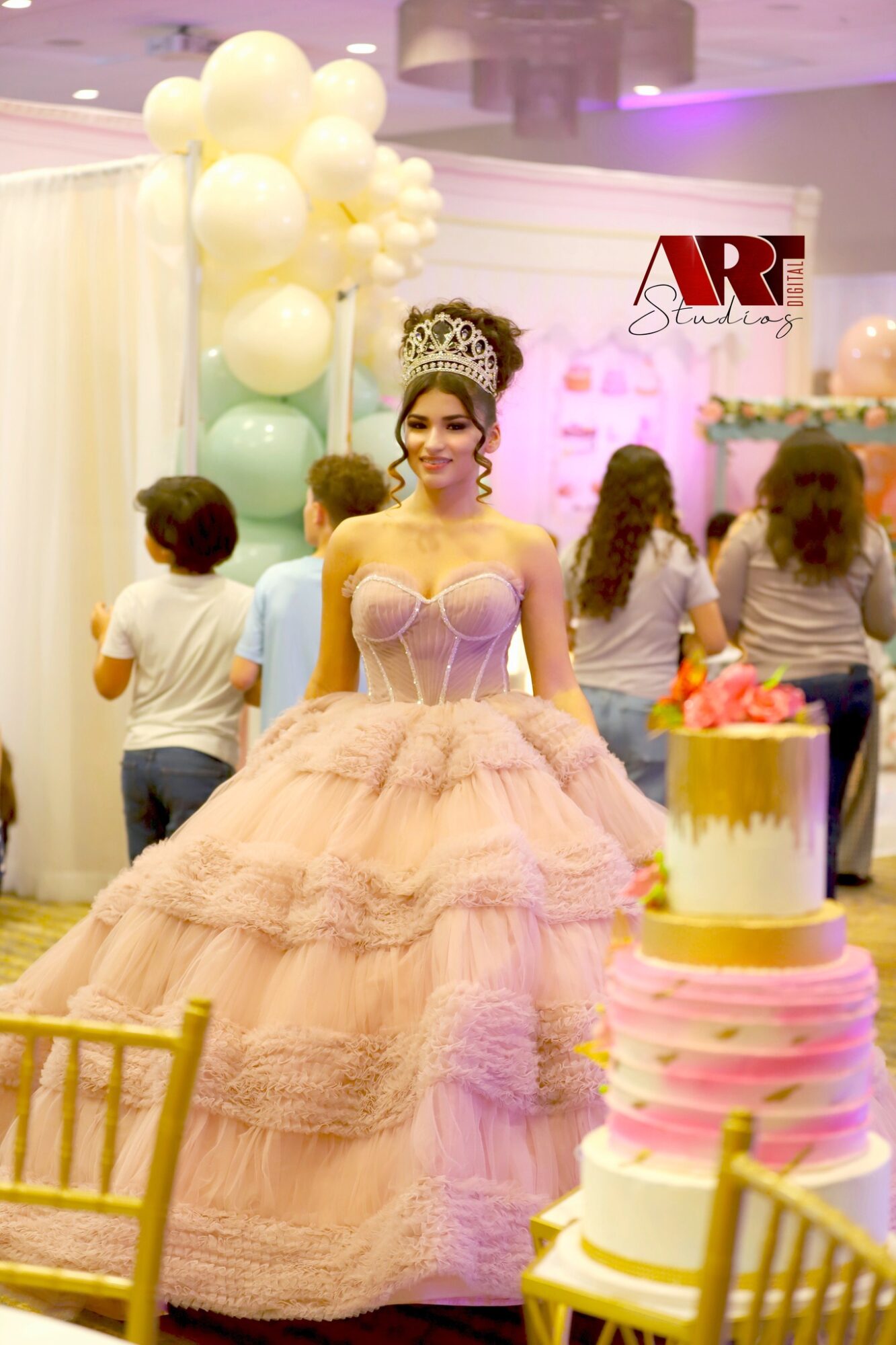 Young woman in a pink ball gown and tiara at a celebration, with balloons and a cake nearby.