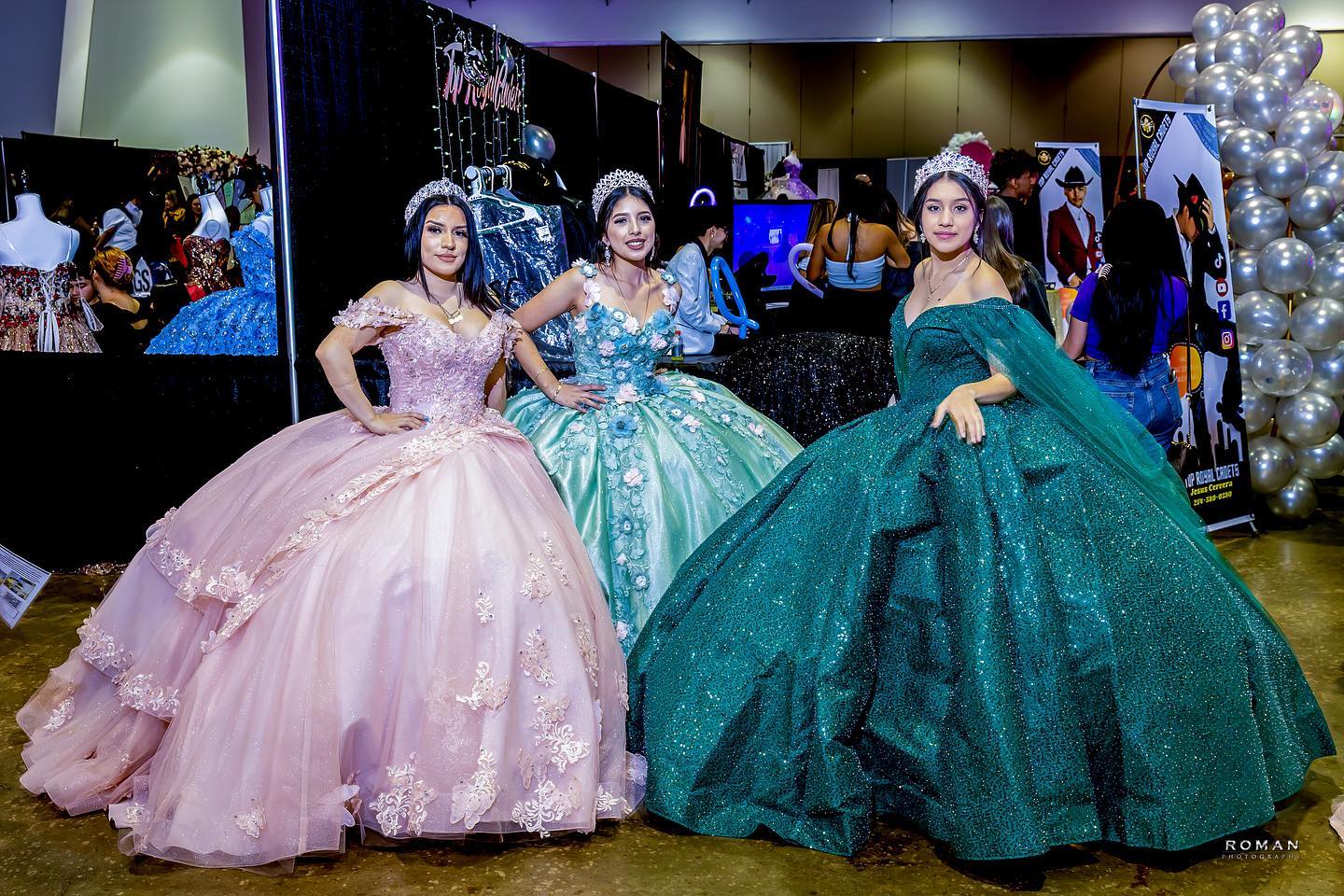 Three women in elaborate ball gowns pose indoors, with balloons and mirrors in the background.