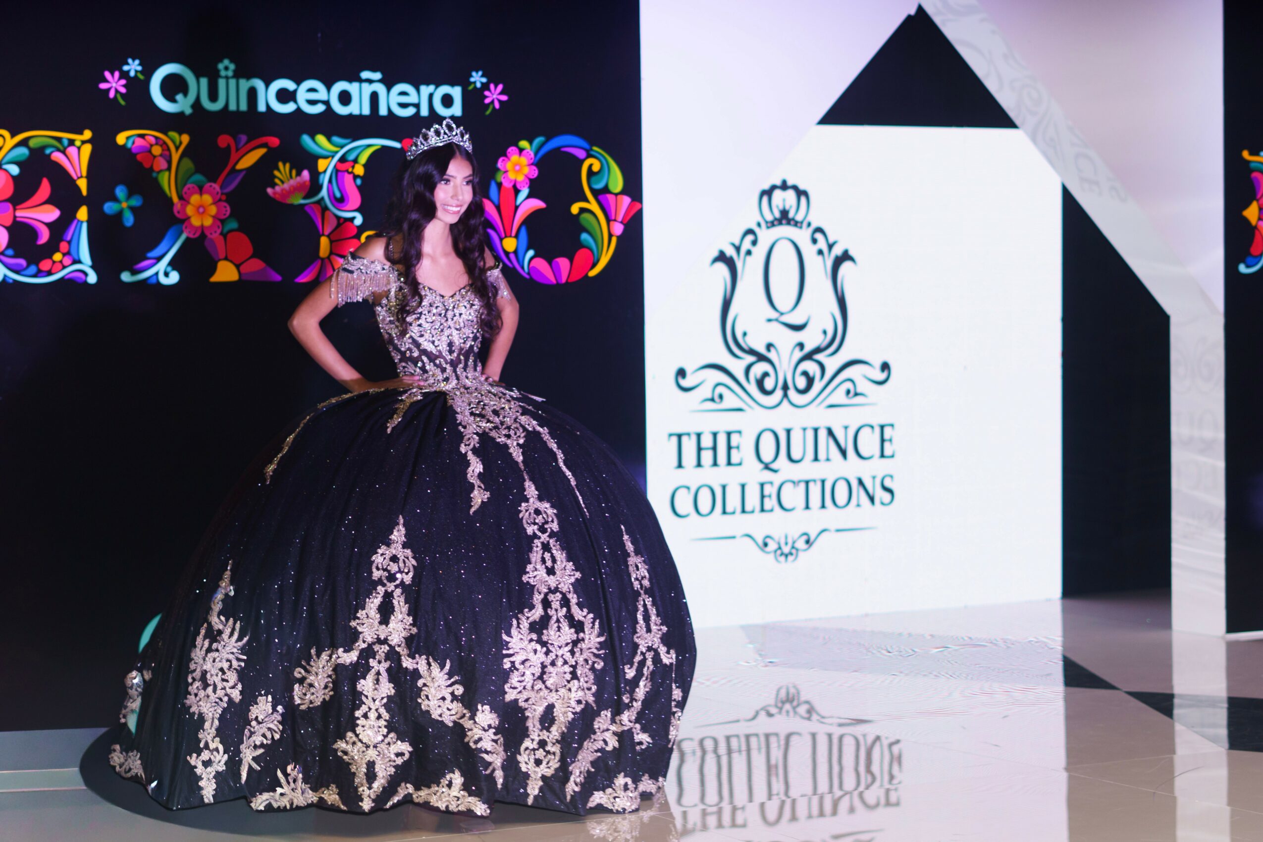 Woman in a black and white gown with floral patterns, standing on stage with event banners and logo behind her.