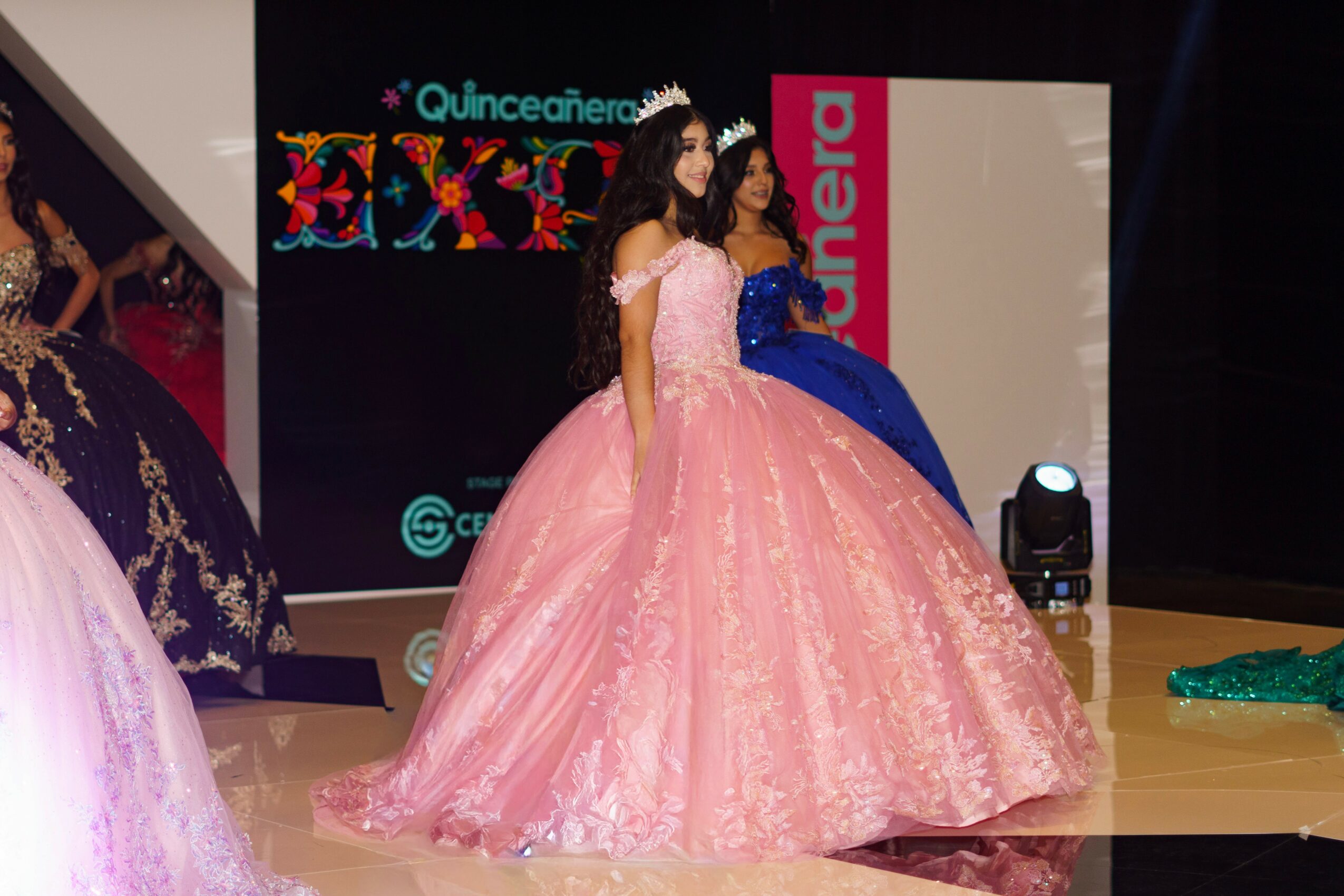 Two women in pink ball gowns and tiaras on stage, with a black background and colorful decorations.