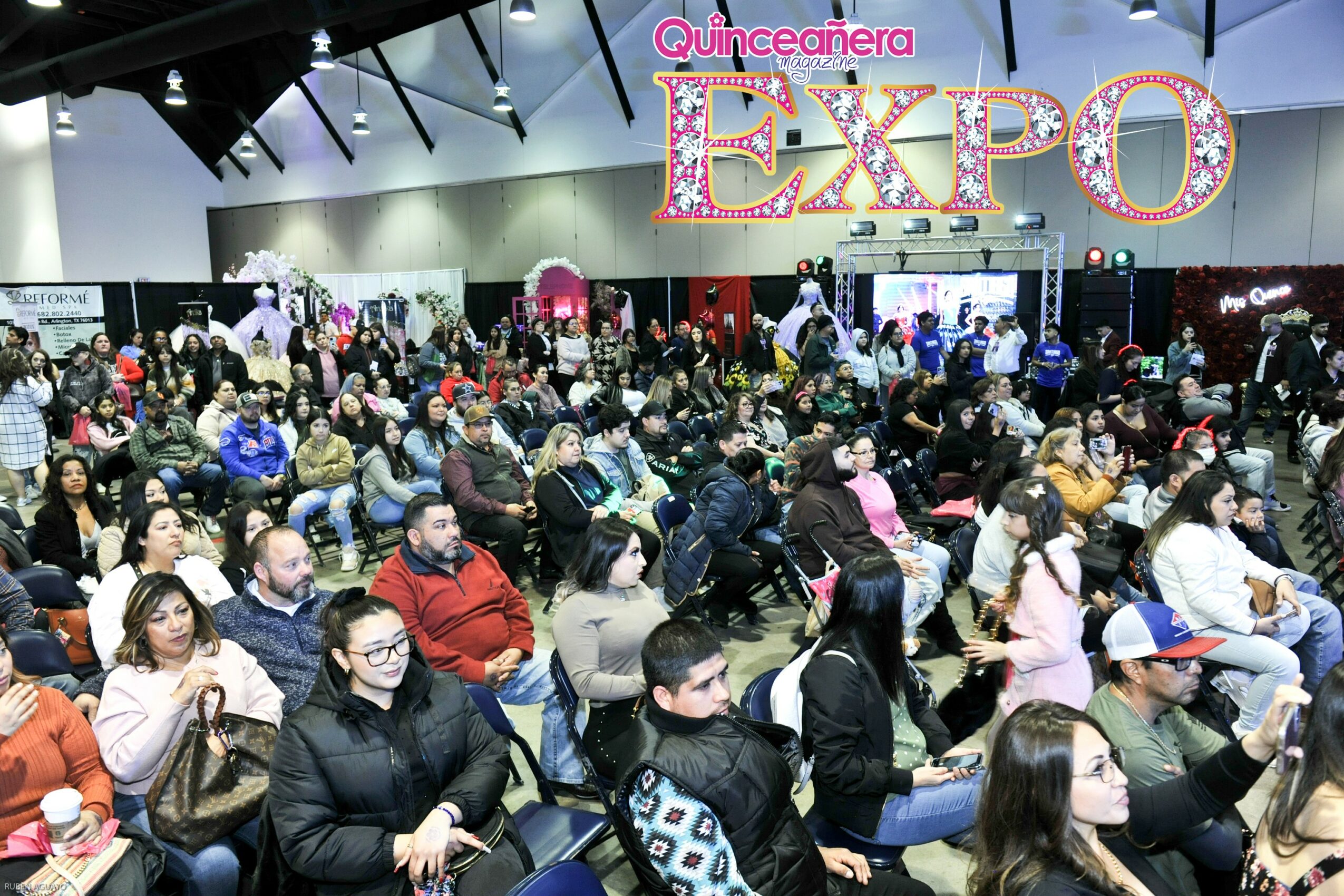 Large crowd seated in an indoor event space with a sign reading 'Quinceañera Expo' overhead.
