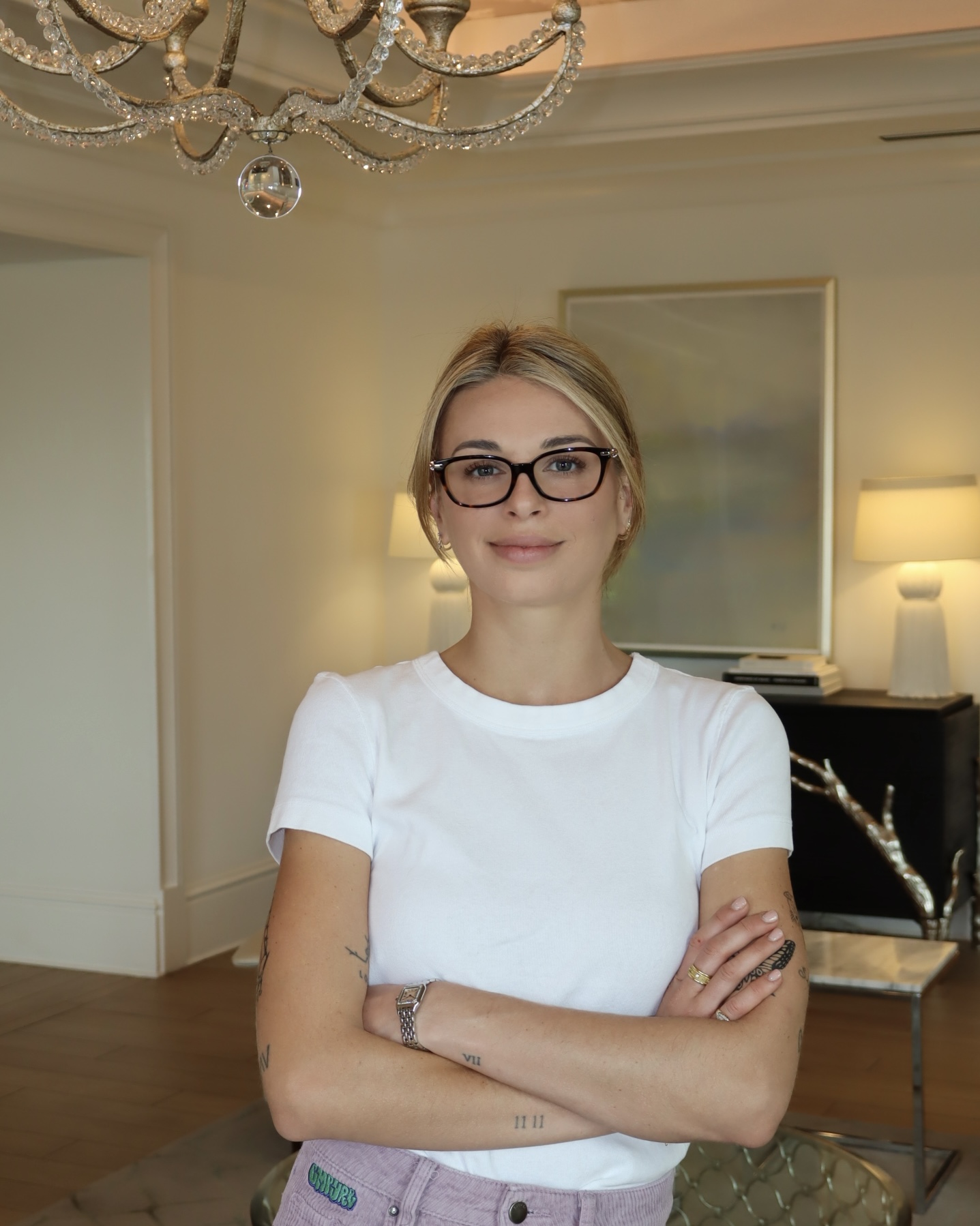 Woman with glasses and blonde hair standing with arms crossed in a living room with chandelier and artwork.