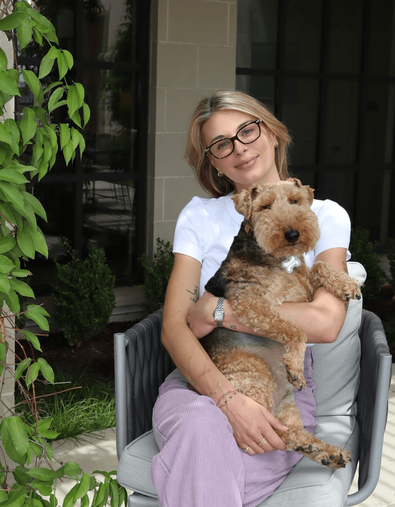 Woman with glasses holding a small dog outdoors near a building and greenery.