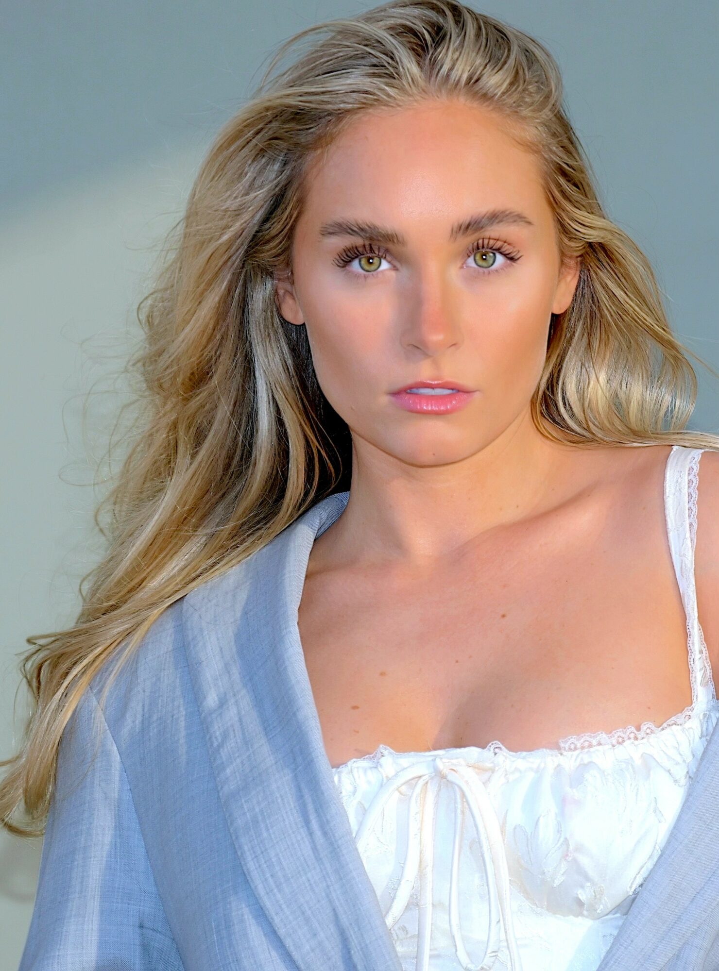 Young woman with long blonde hair, blue eyes, wearing a white top and light blue shawl, looking at camera.