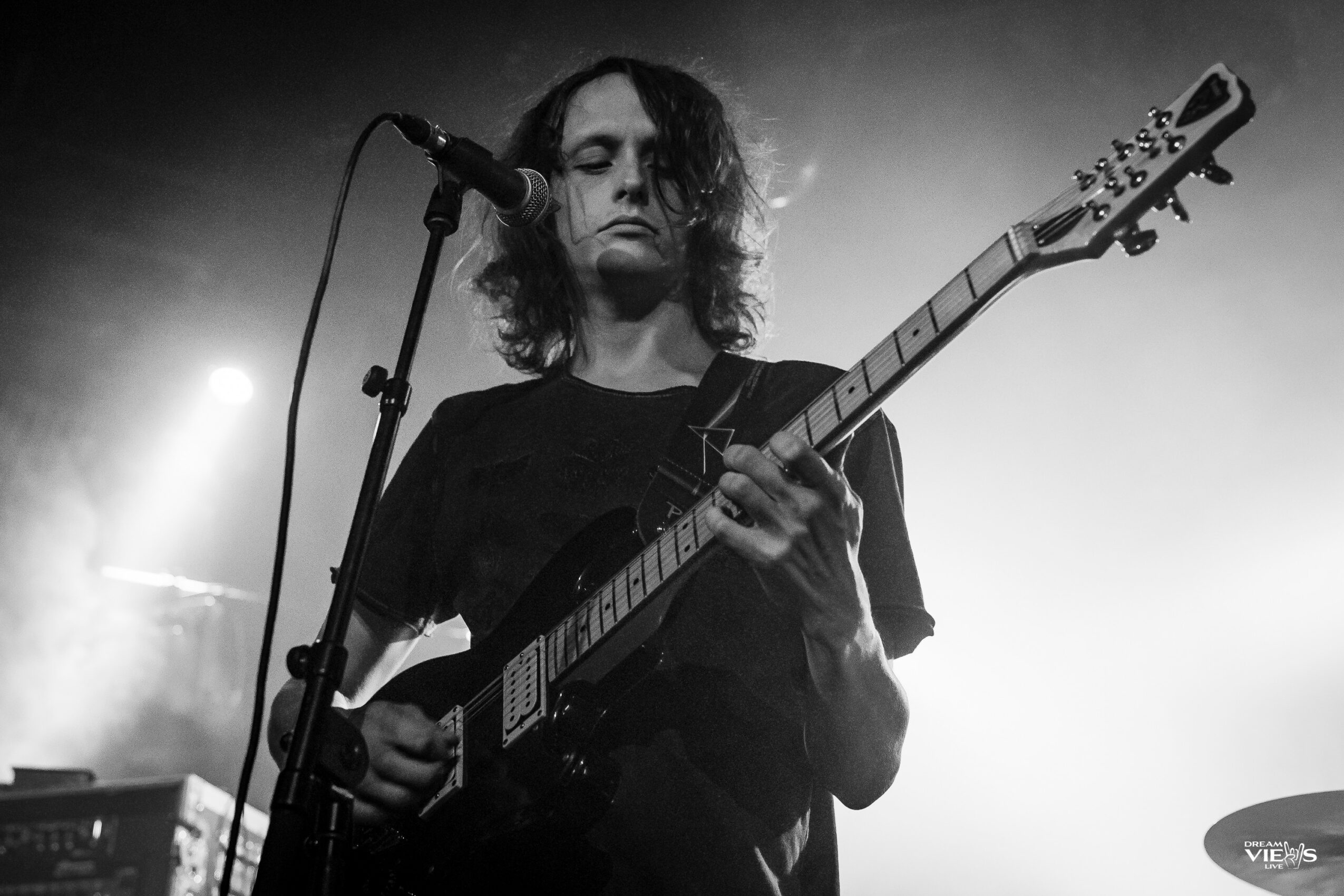 Person with curly hair playing an electric guitar on stage with microphone, black and white image.