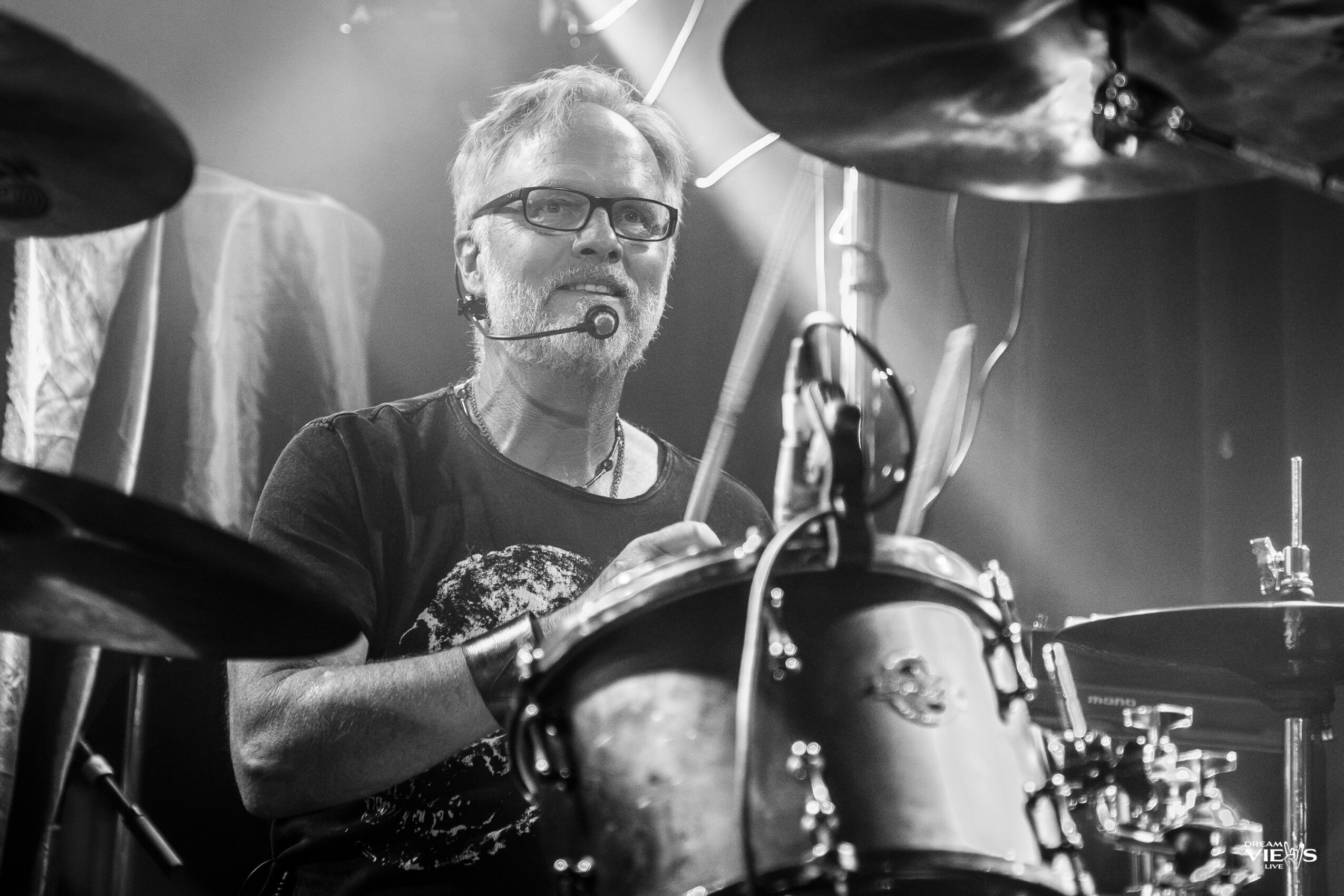 Person playing drums with drumsticks, wearing glasses and a headset, in a performance setting.