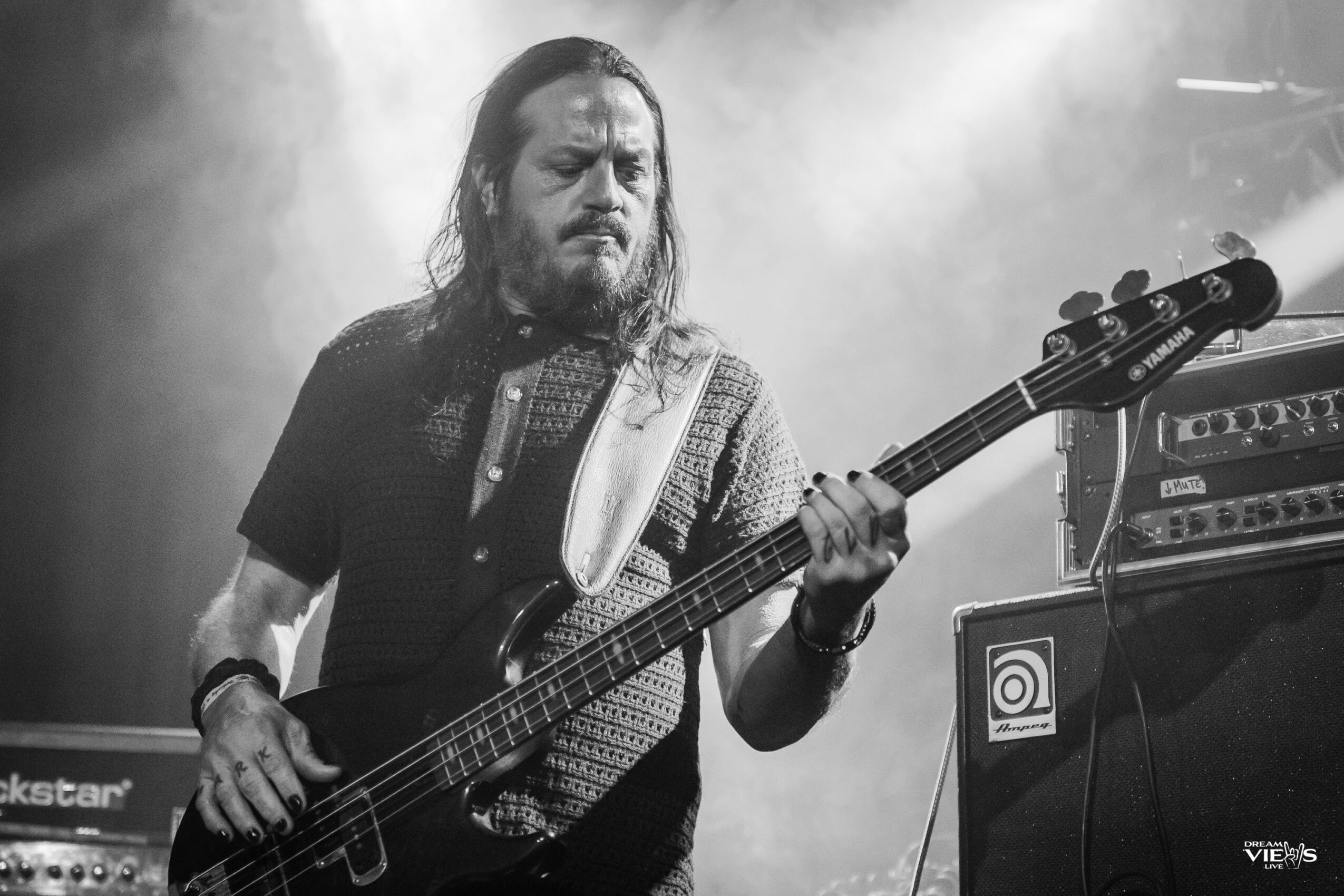 Man with long hair and beard playing bass guitar on stage with equipment in background.