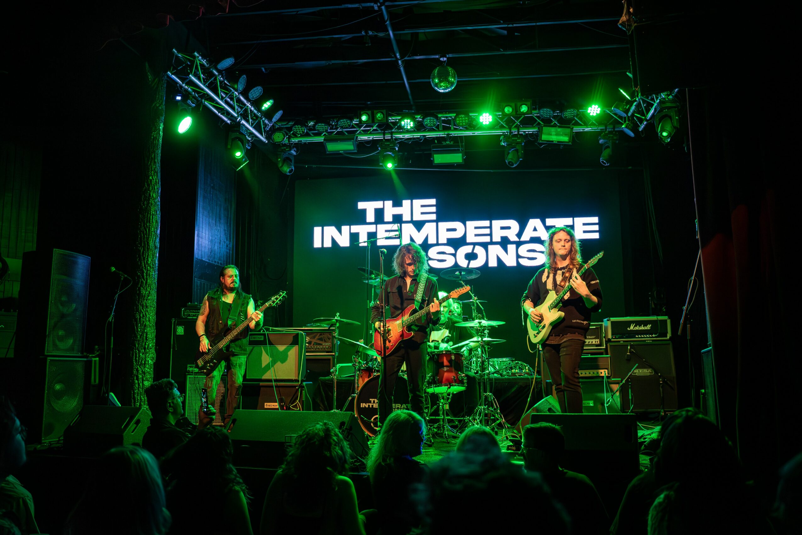 Band performing on stage with green lighting and a large screen displaying 'THE INTERMEDIATE SONS'. Audience visible in foreground.