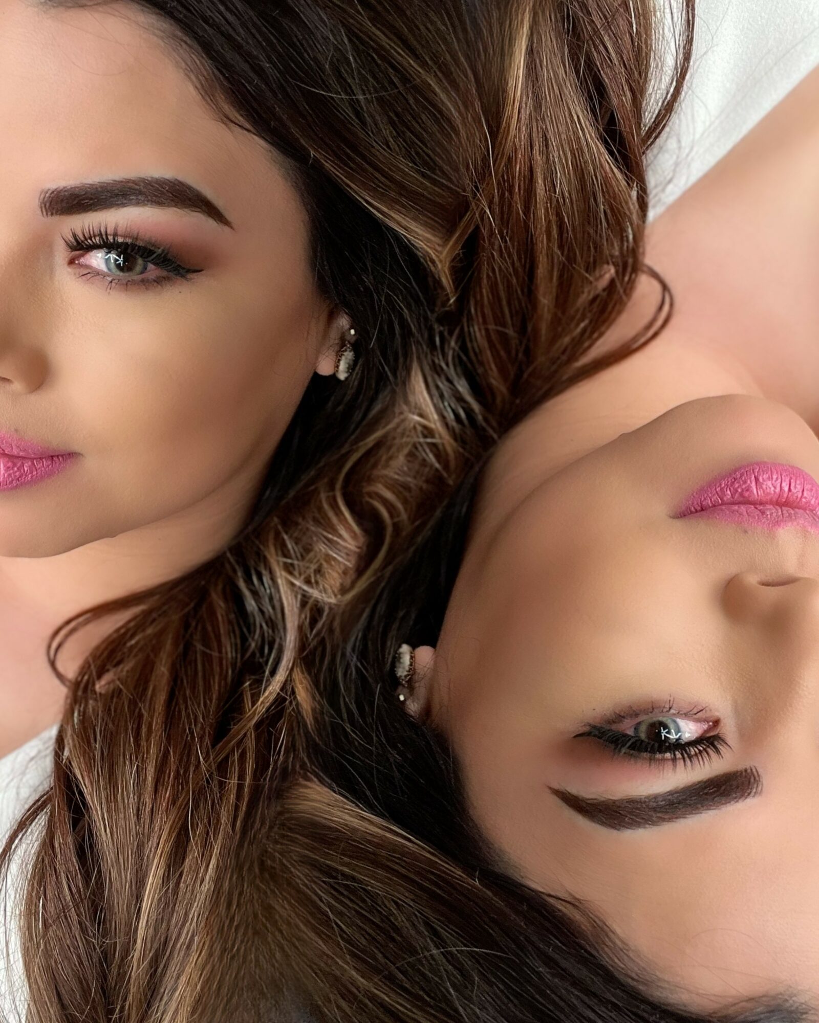 Two women with makeup and long hair lie close, one with eyes open, the other with eyes closed.