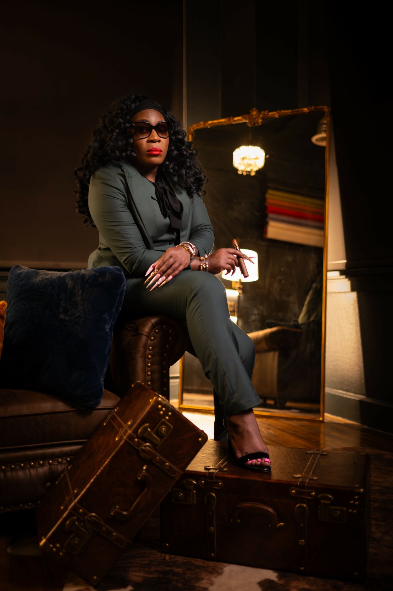 Woman with curly hair and sunglasses sitting on a chair, holding a glass, in a dimly lit room with a mirror.