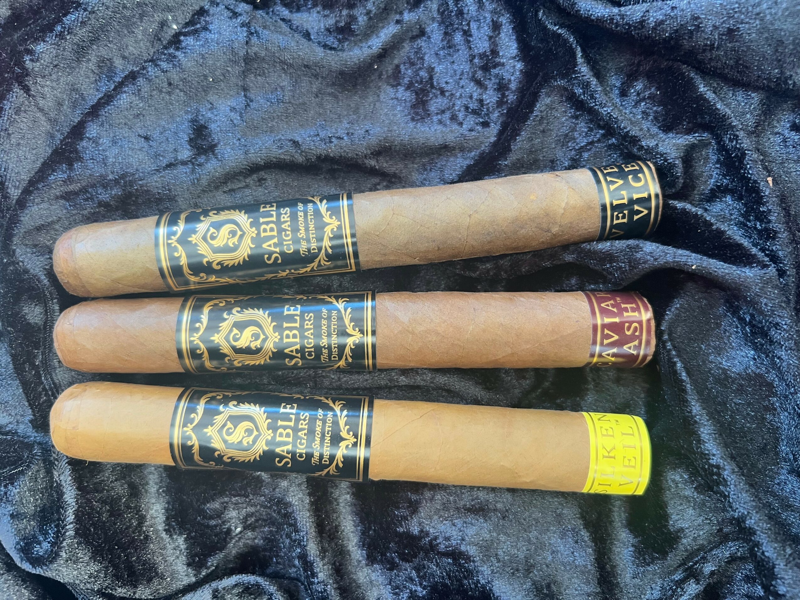 Three cigars with black and gold bands and one with a yellow band, laid on dark fabric.