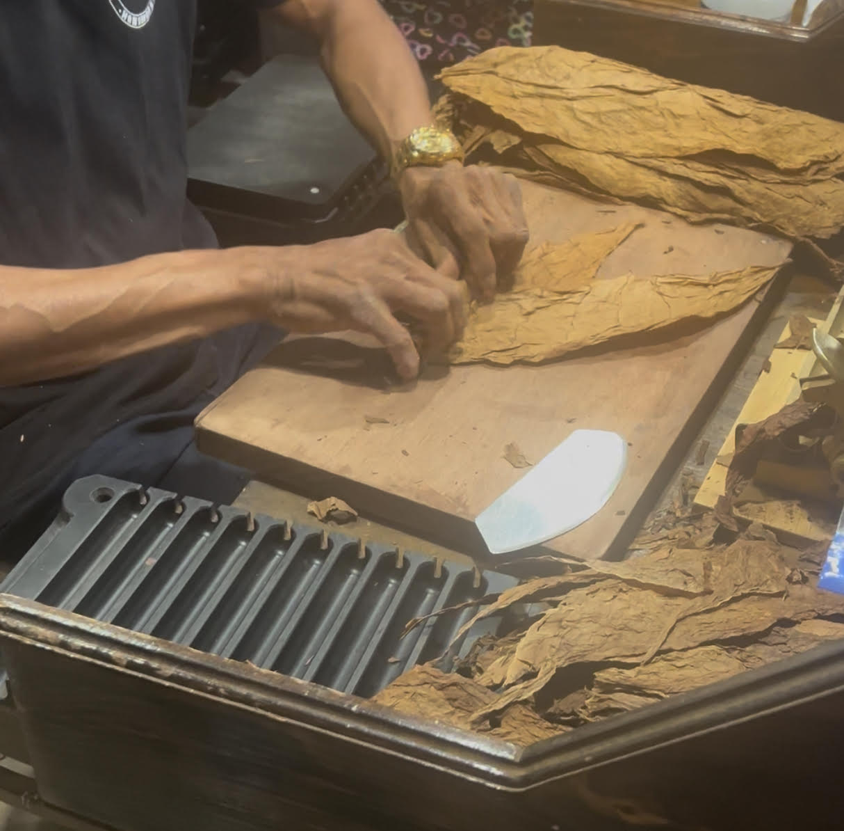 Person's hands working with large sheets of tobacco on a wooden surface, with more sheets nearby.