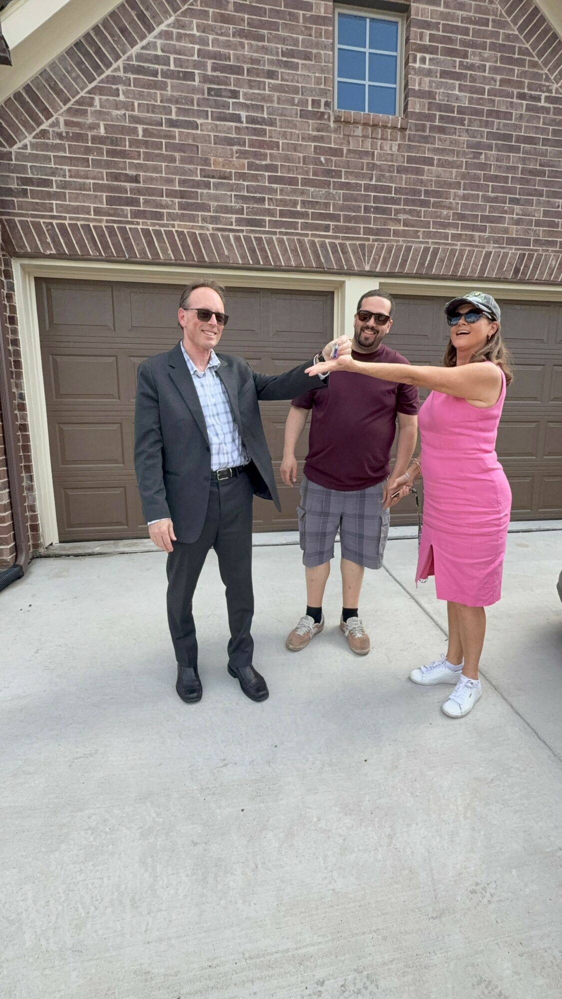 Three people standing outside a brick house, two men and one woman, with the woman and one man holding each other's shoulders.