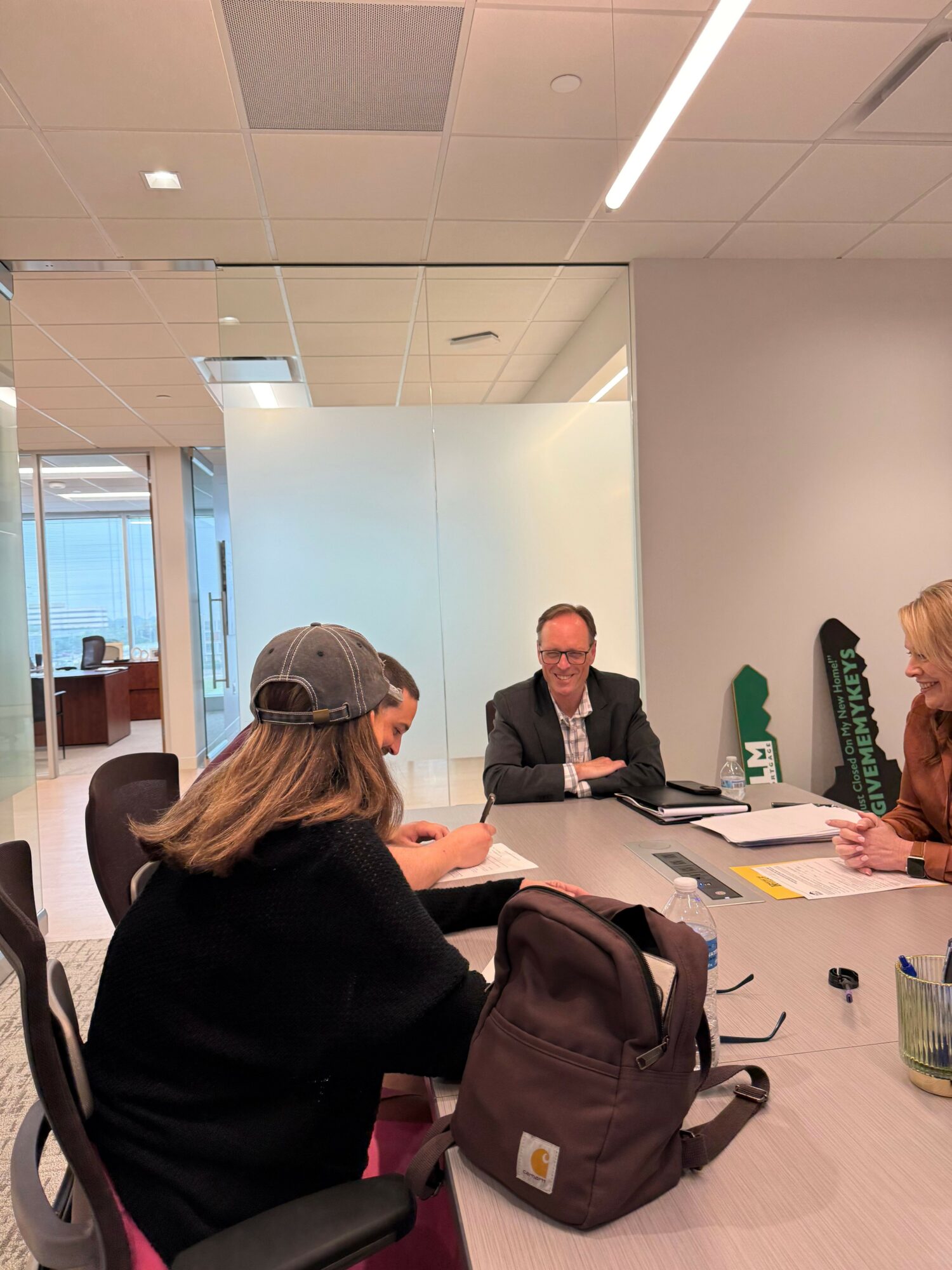 Four people seated around a conference table in a modern office, engaged in discussion, with documents and a backpack on the table.