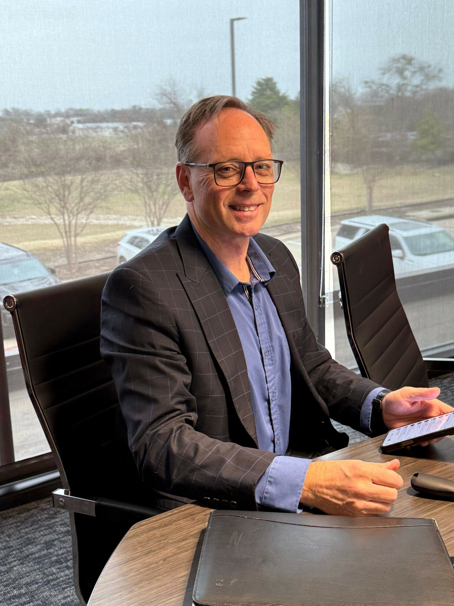 Man with glasses in a suit sitting at a conference table near a window, smiling, with cars outside.