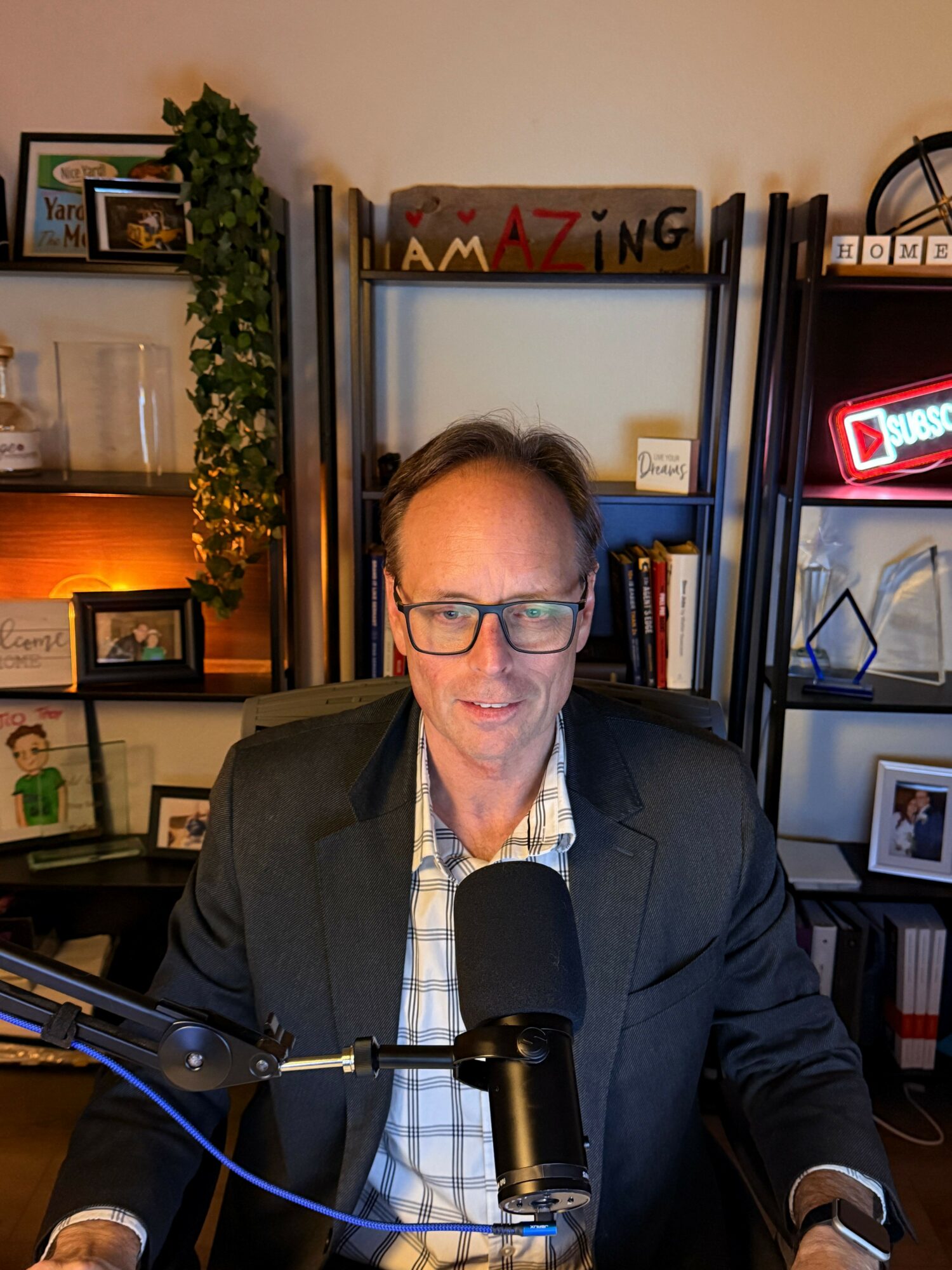 Man with glasses in a suit sitting at a desk with a microphone, bookshelf behind him, indoor setting.