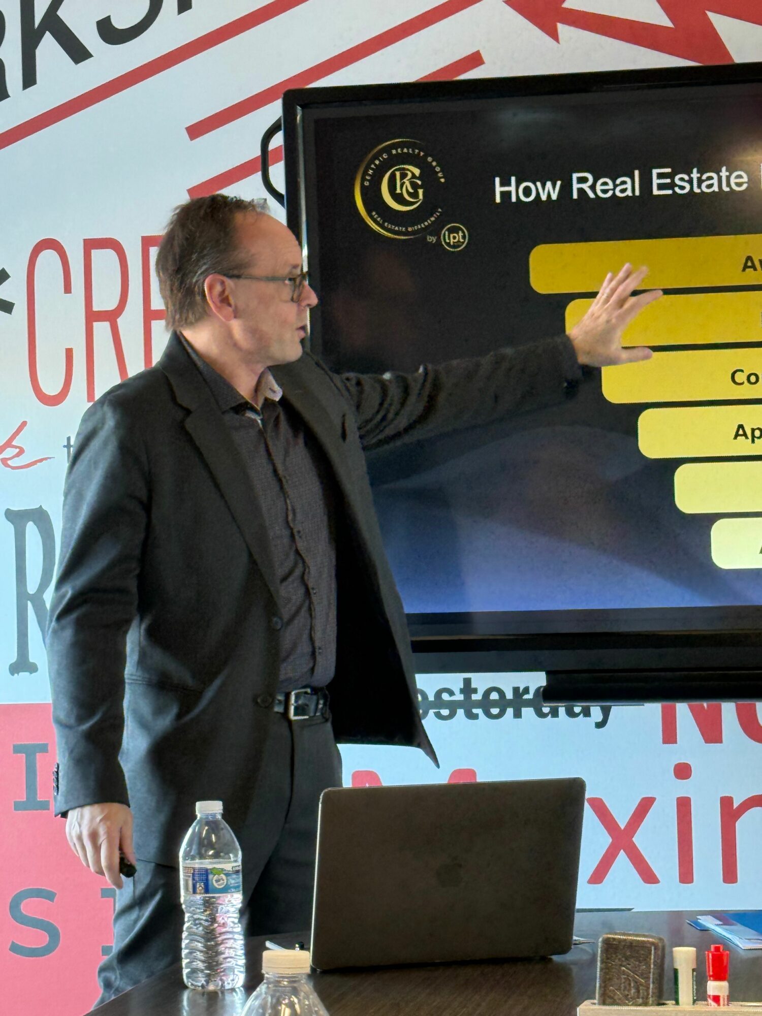 Man in dark suit and glasses points at a presentation slide on a screen, with a laptop and water bottle on table.