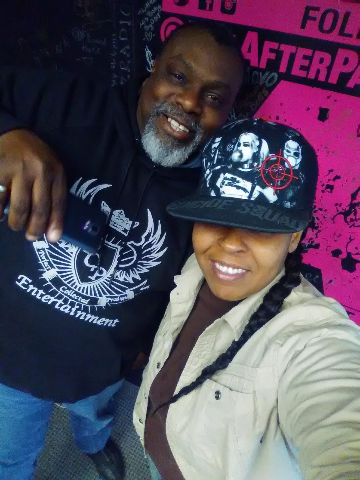 Two people smiling, one wearing a black cap with graphic design, the other in a beige jacket, in front of a pink background.