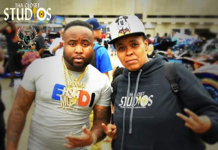 Two people standing outdoors, one wearing a white cap and hoodie, the other in a gray shirt with gold chains.